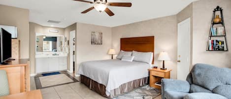 A bedroom with a bed, bedside lamps, a ceiling fan, a bookshelf, and a view into the bathroom.