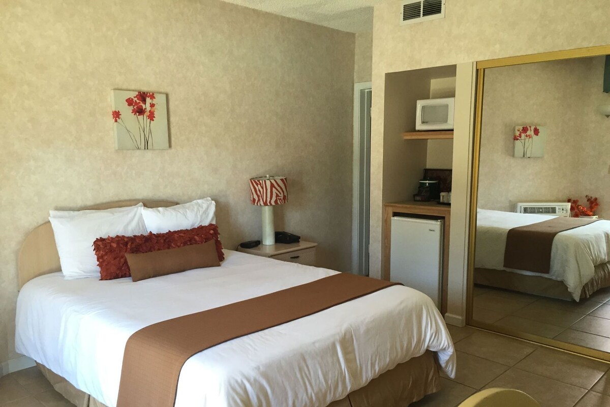 A hotel room with a bed, a bedside table with a lamp, a small fridge, a microwave, and a mirror.