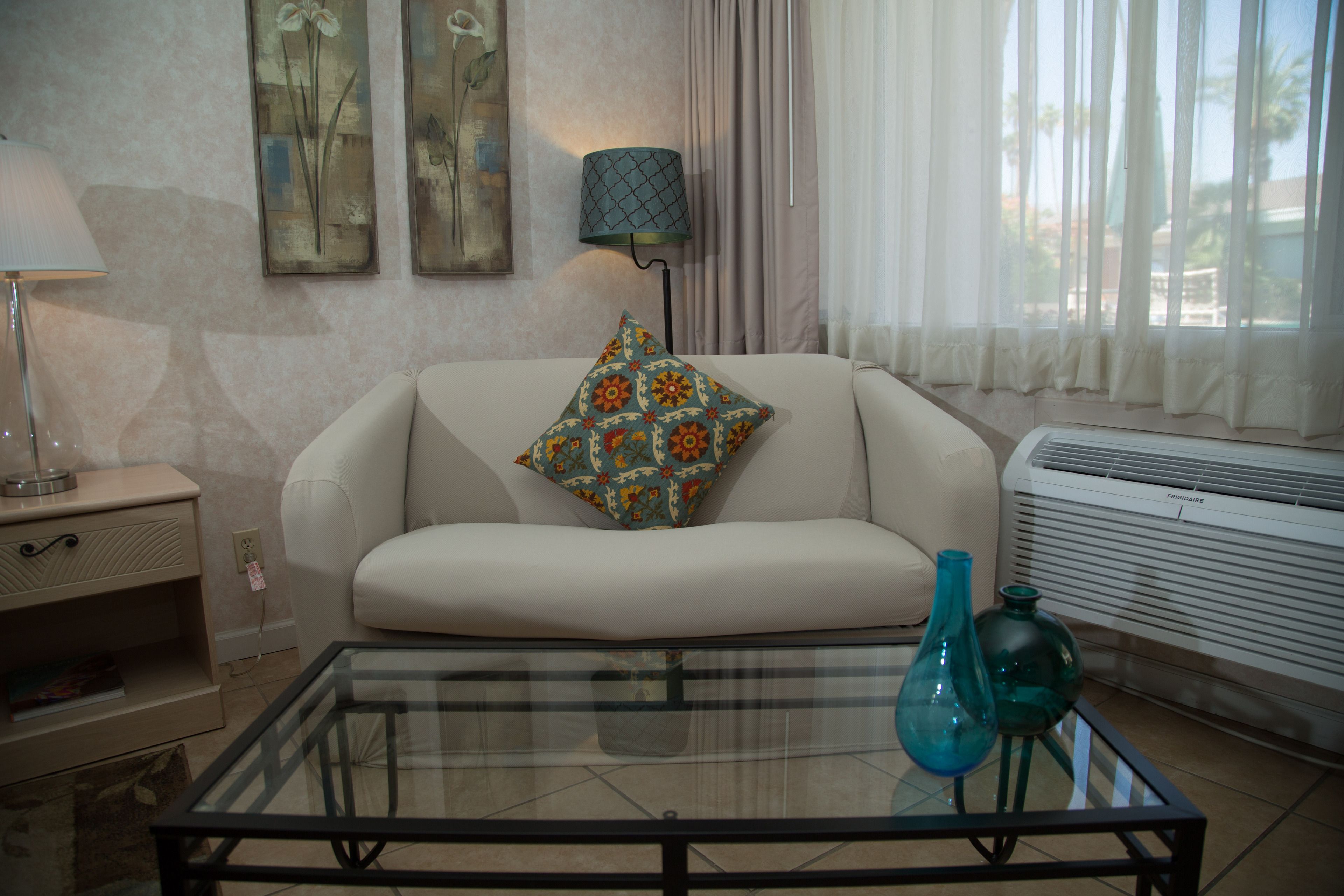 A living room with a sofa, a glass coffee table, a lamp, and a window with curtains.