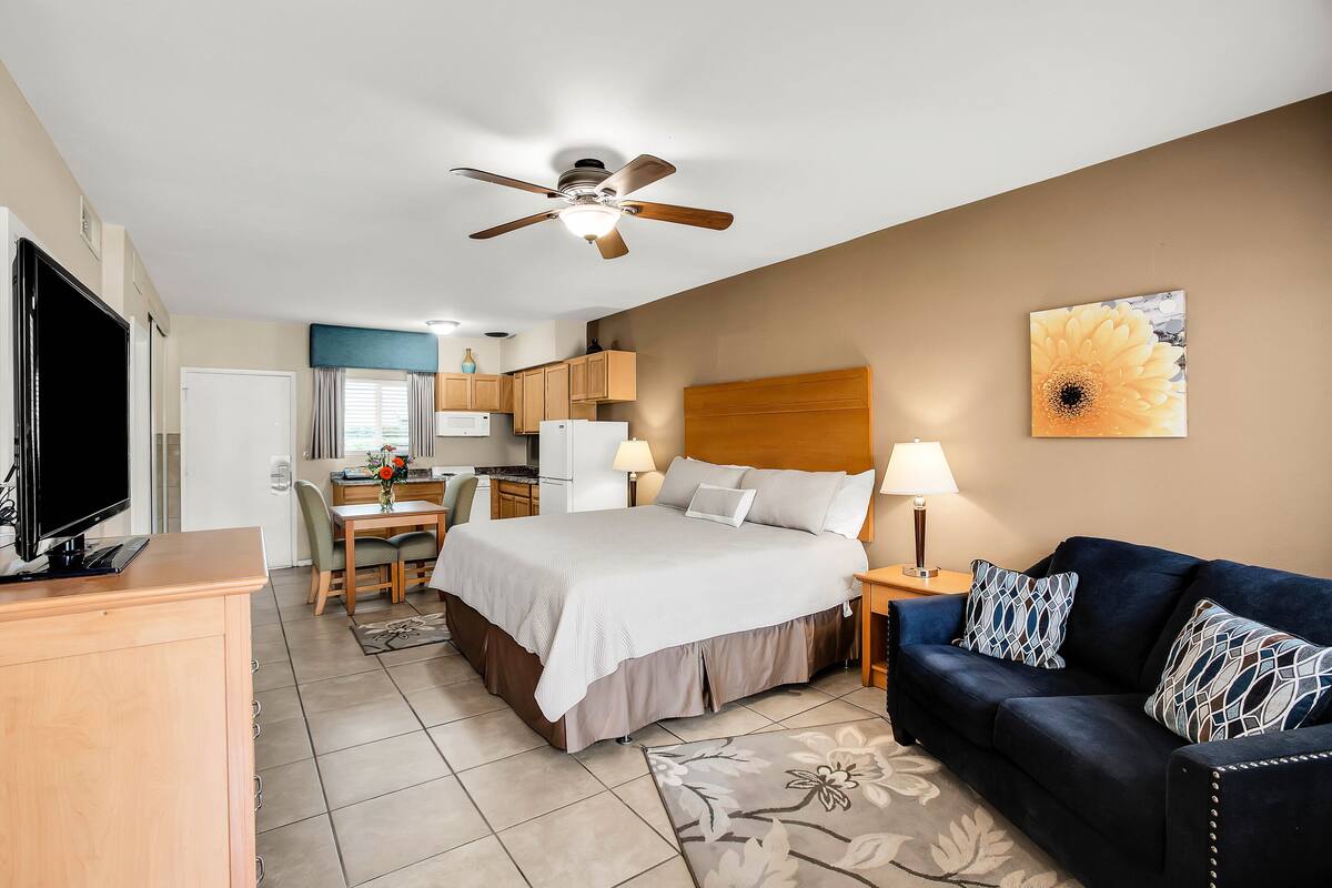 A hotel room with a bed, a sofa, a TV, a ceiling fan, a kitchen area, and a dining table.