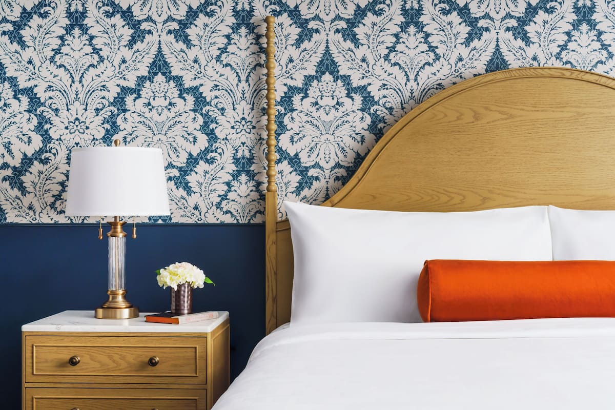 A neatly made bed with white and orange pillows, a wooden headboard, and a bedside table with a lamp and a vase of flowers.