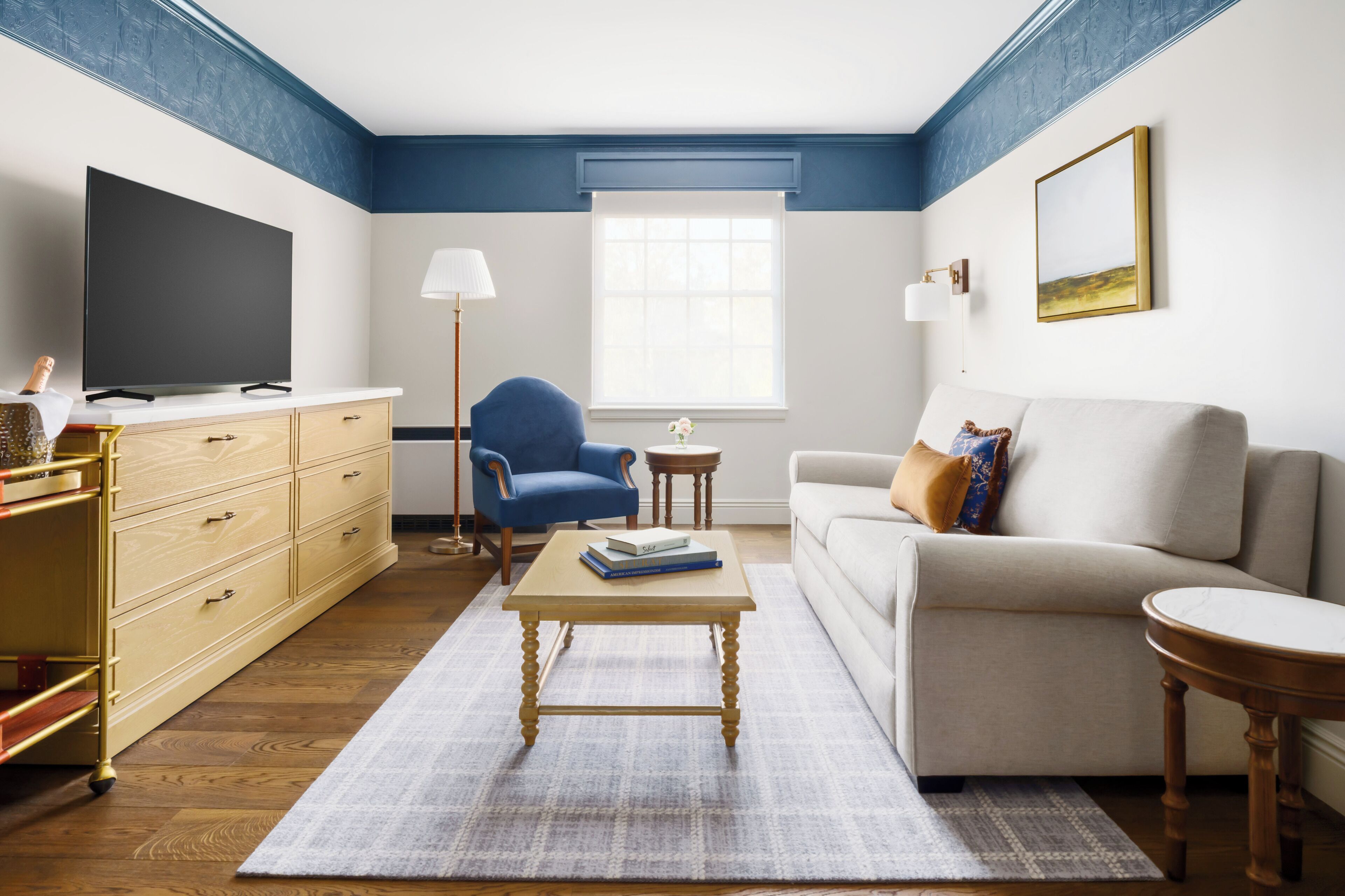 A living room with a flat-screen TV, a blue armchair, a beige sofa, a wooden coffee table, and a small side table with a lamp.