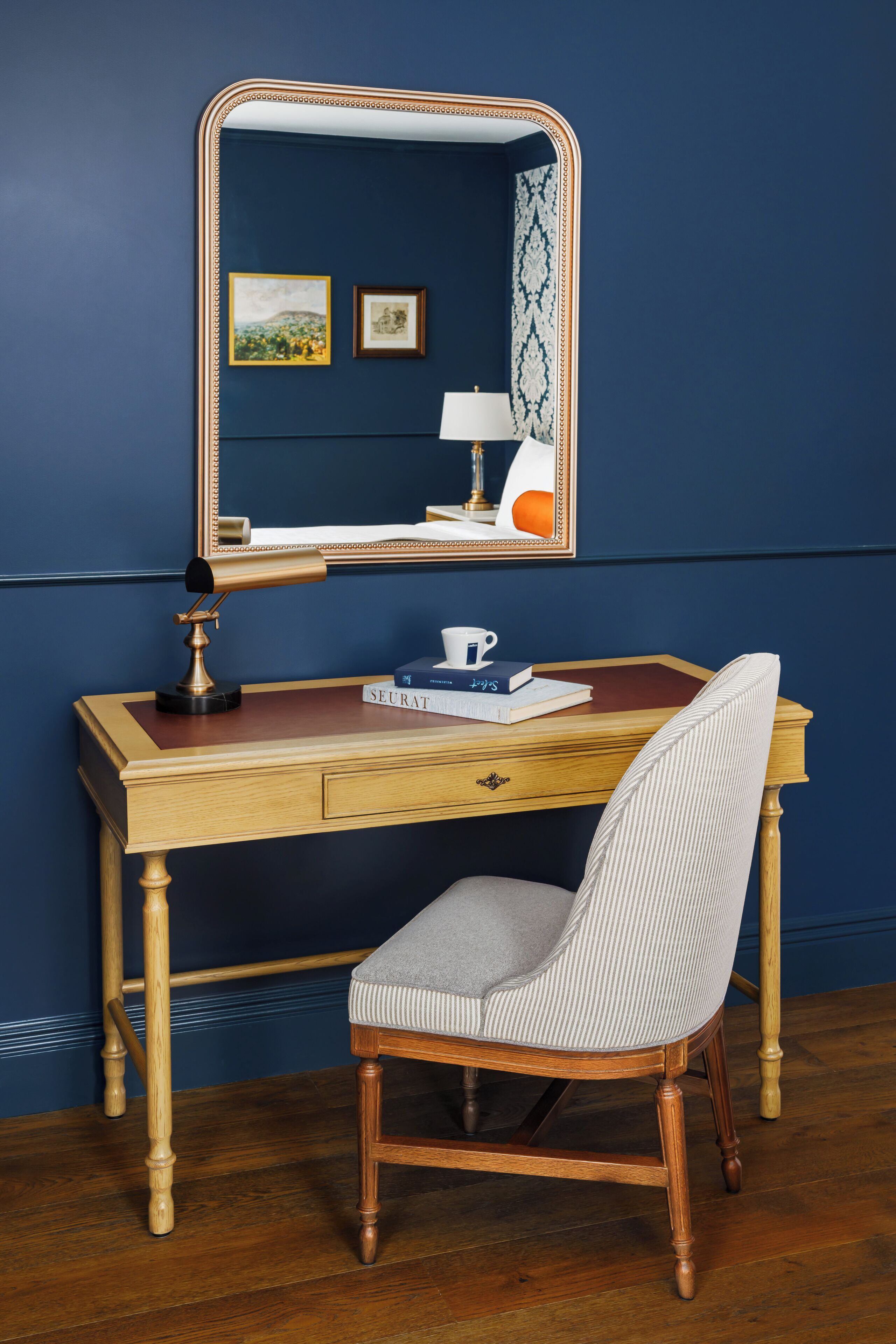 A wooden desk with a mirror, a lamp, and a chair. There are books and a cup on the desk.