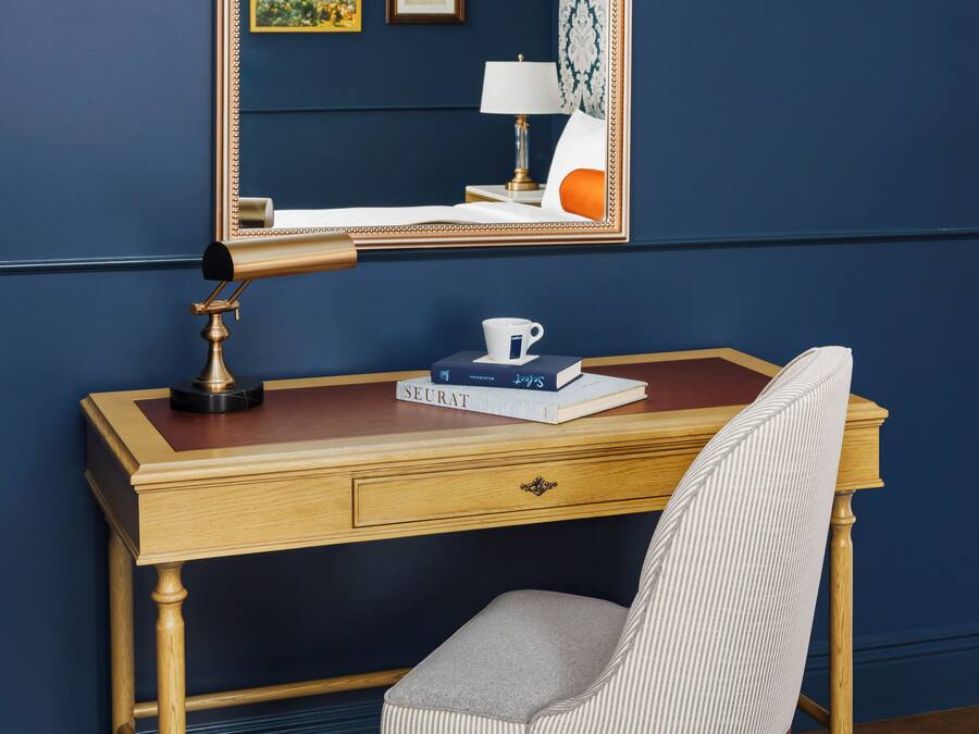 A wooden desk with a mirror, a lamp, and a chair. There are books and a cup on the desk.