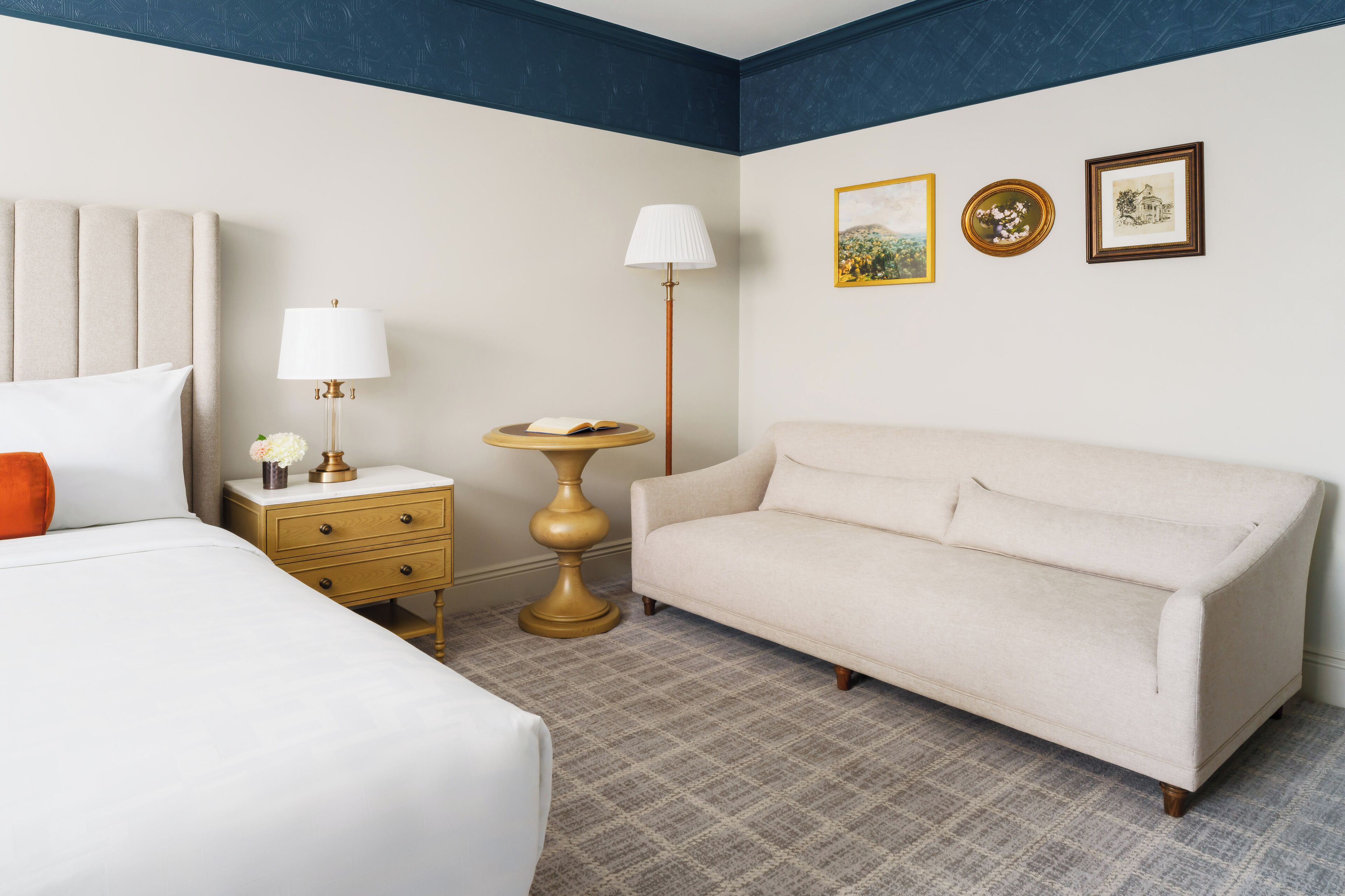 Una habitación de hotel con cama, sofá, mesita de noche con lámpara, una pequeña mesa y cuadros en la pared.