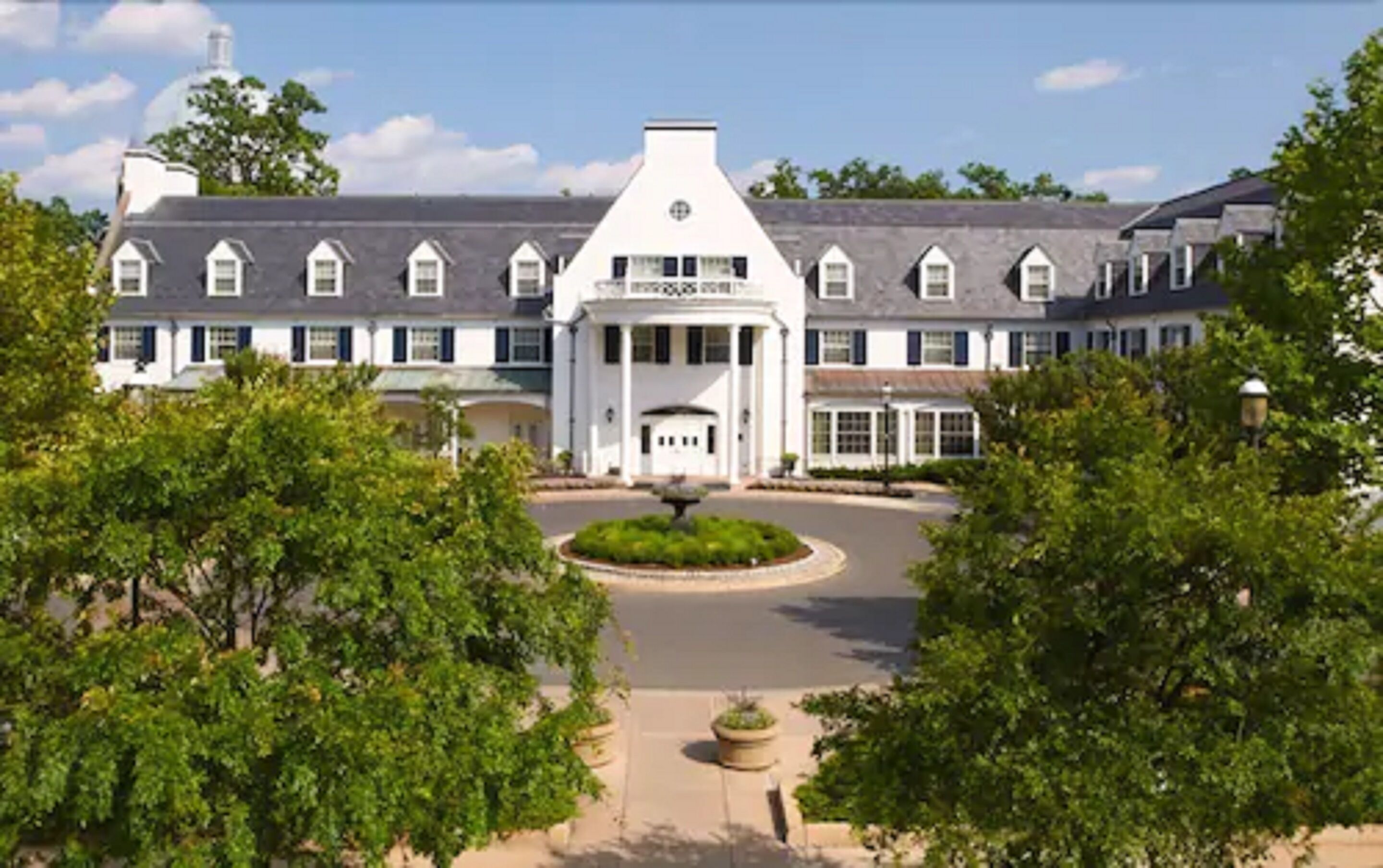 A large, white, two-story building with a symmetrical facade, a central entrance with a pediment, and a landscaped courtyard with a circular garden in the middle.
