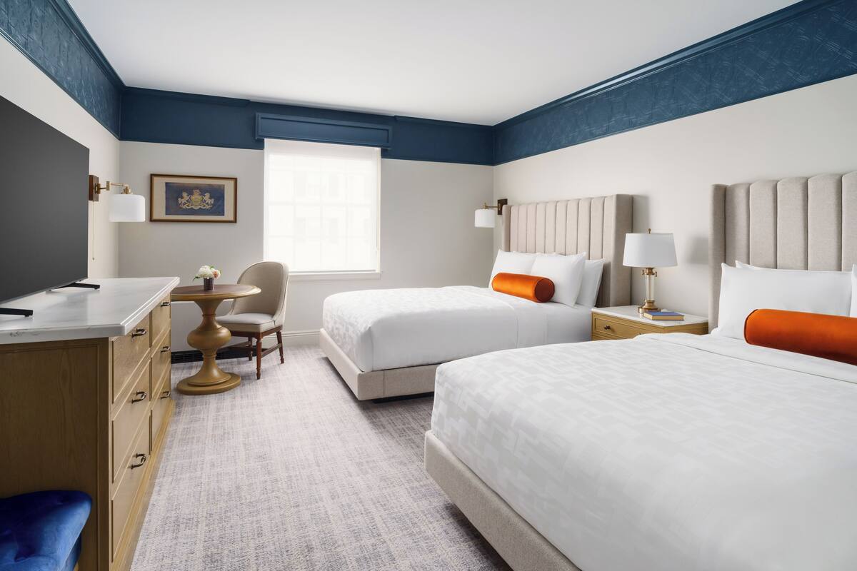 A hotel room with two beds, a desk, a chair, a small table, and a window.