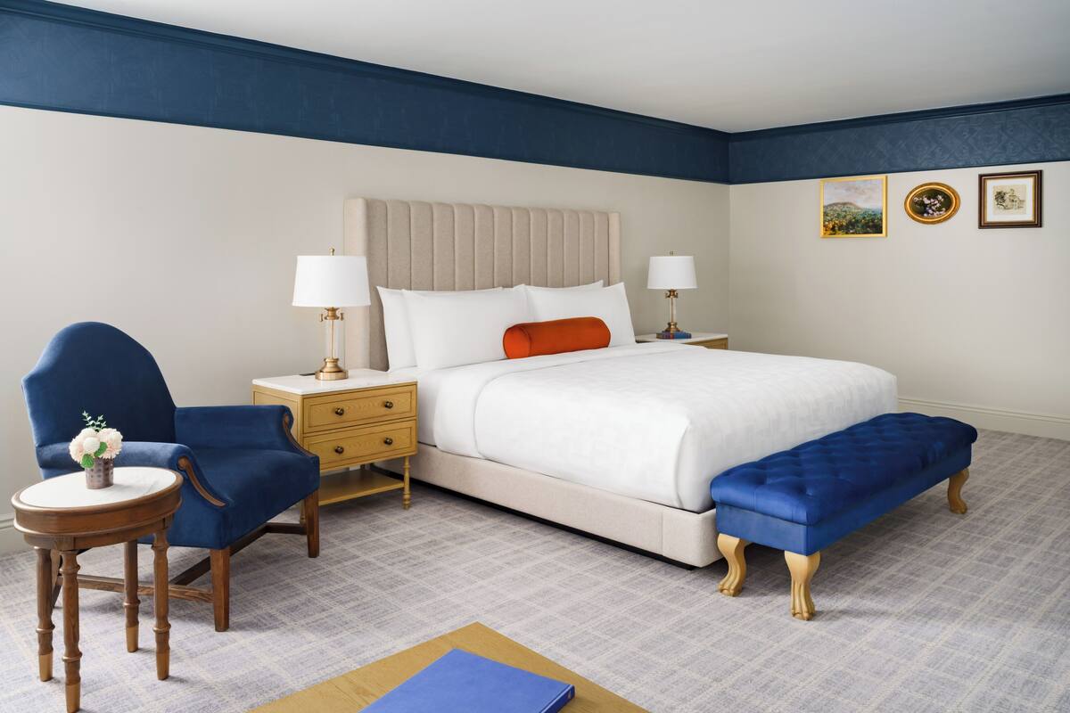 A hotel room with a bed, two nightstands, a blue armchair, a wooden side table, and a bench.