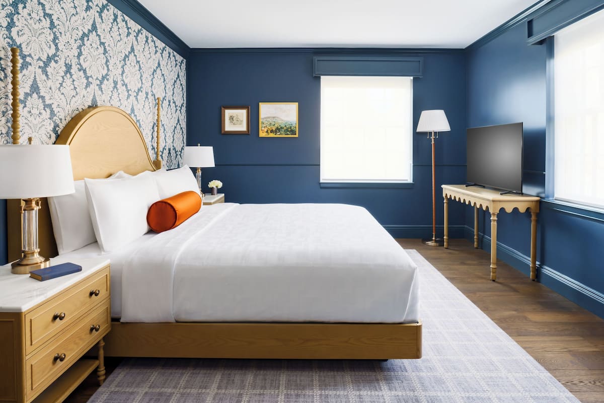 A neatly arranged bedroom with a large bed, bedside lamps, a television on a small table, and a window providing natural light.