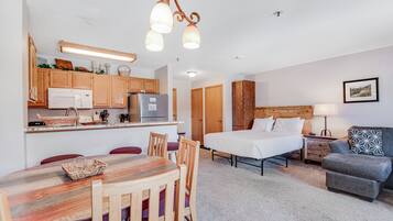 Een compacte hotelkamer met kitchenette, een eethoek met houten tafel en stoelen, een bed met grijs gestoffeerd hoofdeinde, en een grijze bank met gekleurde kussens.