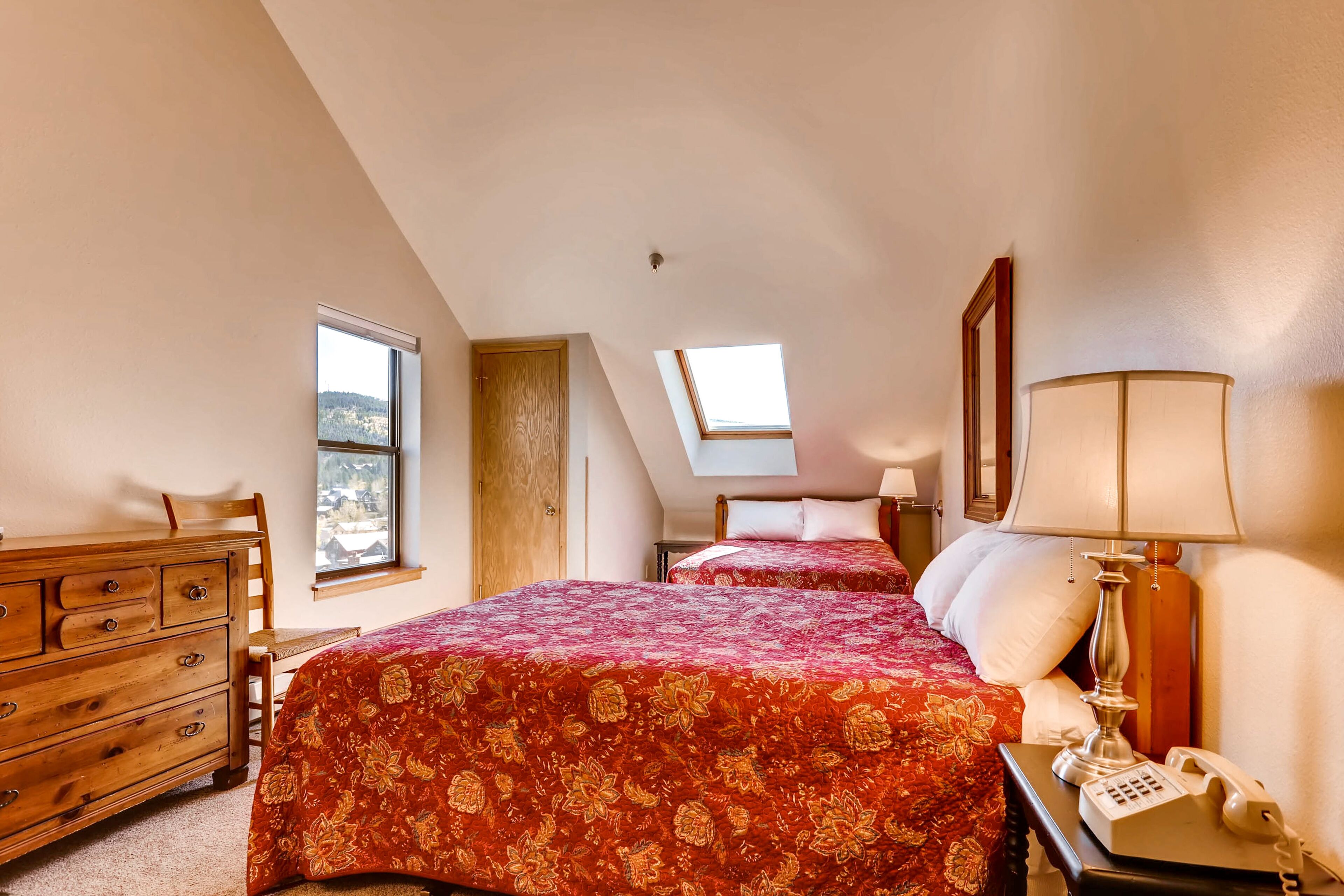 A bedroom with two beds, a wooden dresser, a nightstand with a lamp, and a window with a view.