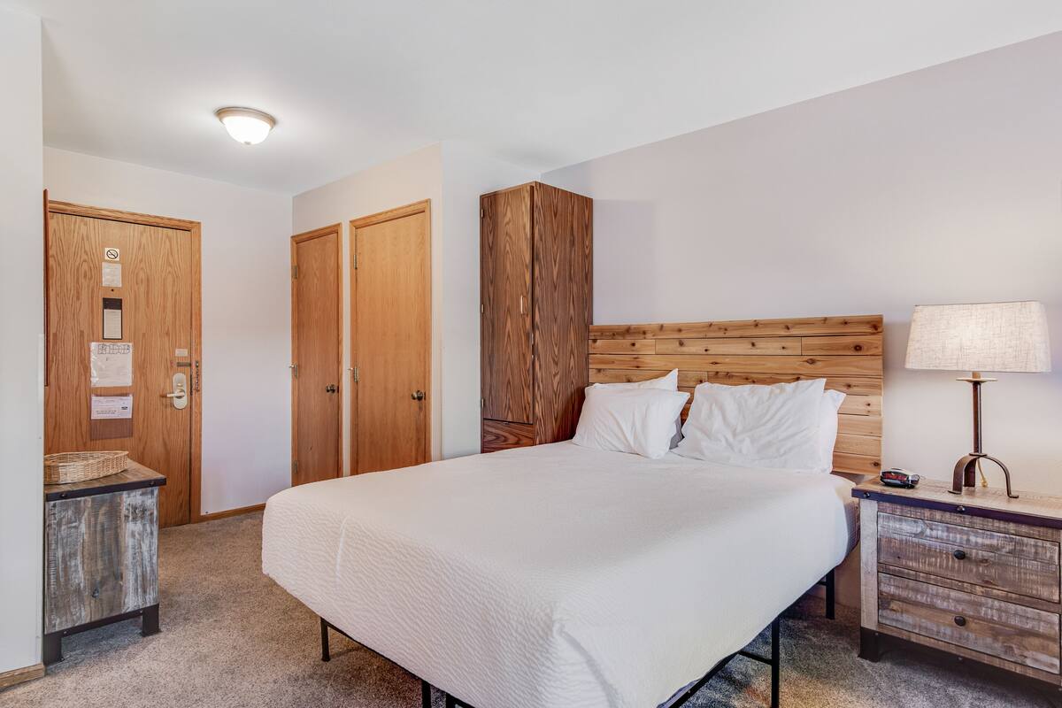 A neatly arranged bedroom with a large bed, wooden wardrobe, and a bedside table with a lamp.
