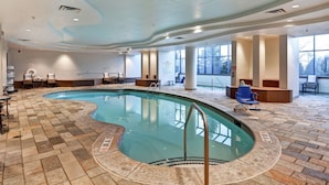 Une piscine intérieure dotée d’un plafond aux lignes ondulantes, d’un sol carrelé et d’un espace aménagé au bord de la piscine avec des chaises et des tables.