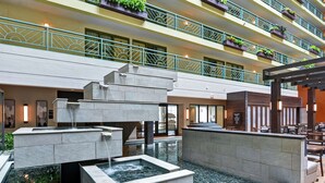 Un hall d’hôtel à plusieurs étages, doté d’un élément décoratif central en eau, d’un espace salon extérieur et de balcons agrémentés de plantes.
