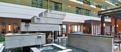 A multi-story hotel lobby with a central water feature, outdoor seating, and balconies with greenery.