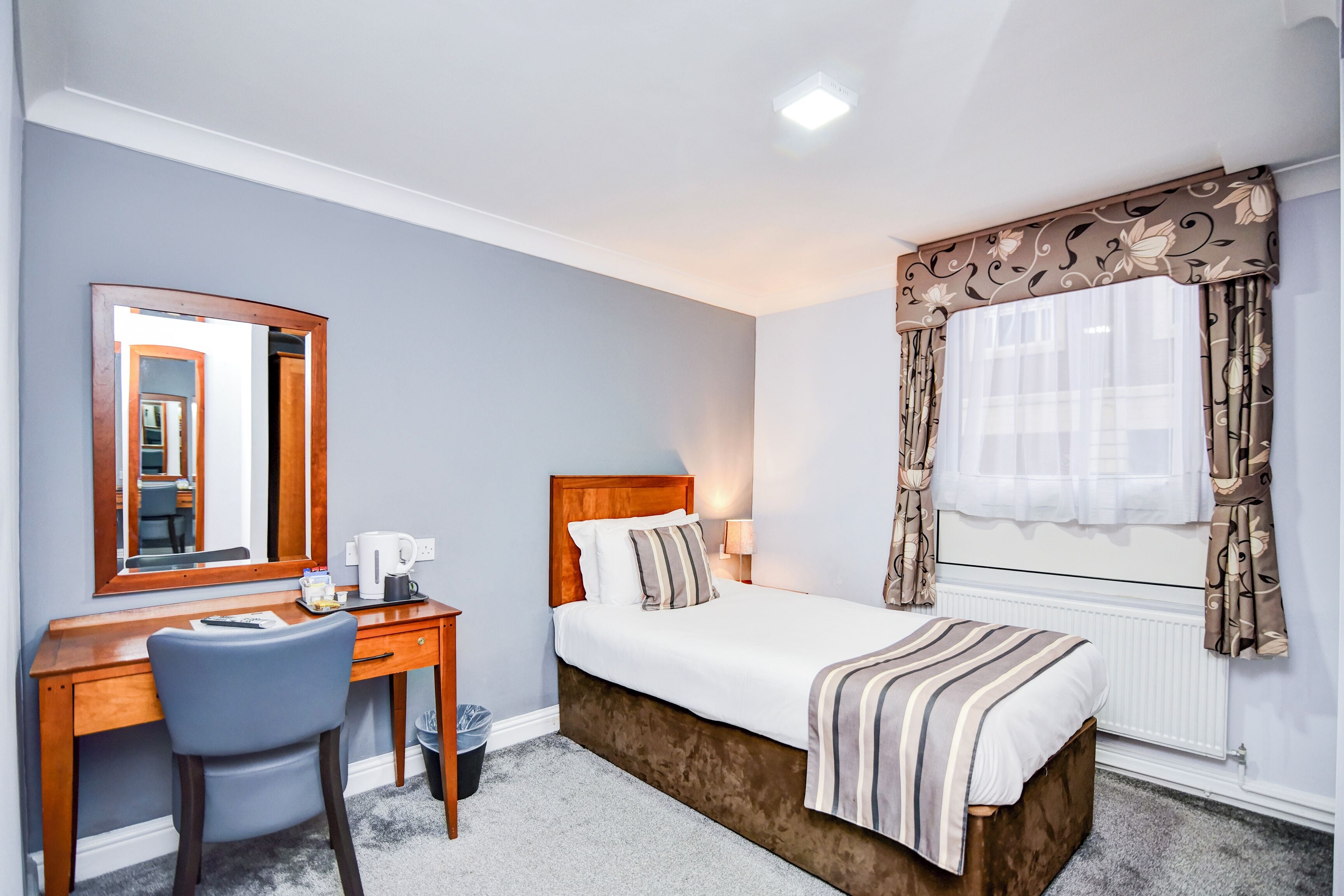 Una habitación de hotel con un escritorio de madera, una silla, una cama con manta a rayas, un espejo grande y cortinas con motivos florales.