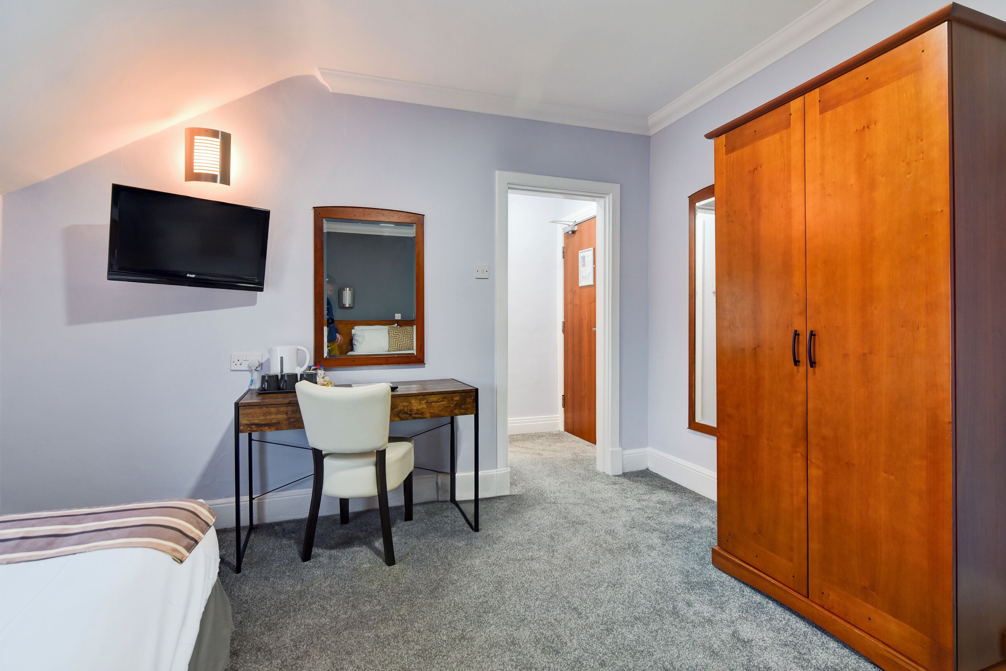 A hotel room with a bed, a desk with a chair, a television, a wardrobe, and a mirror.