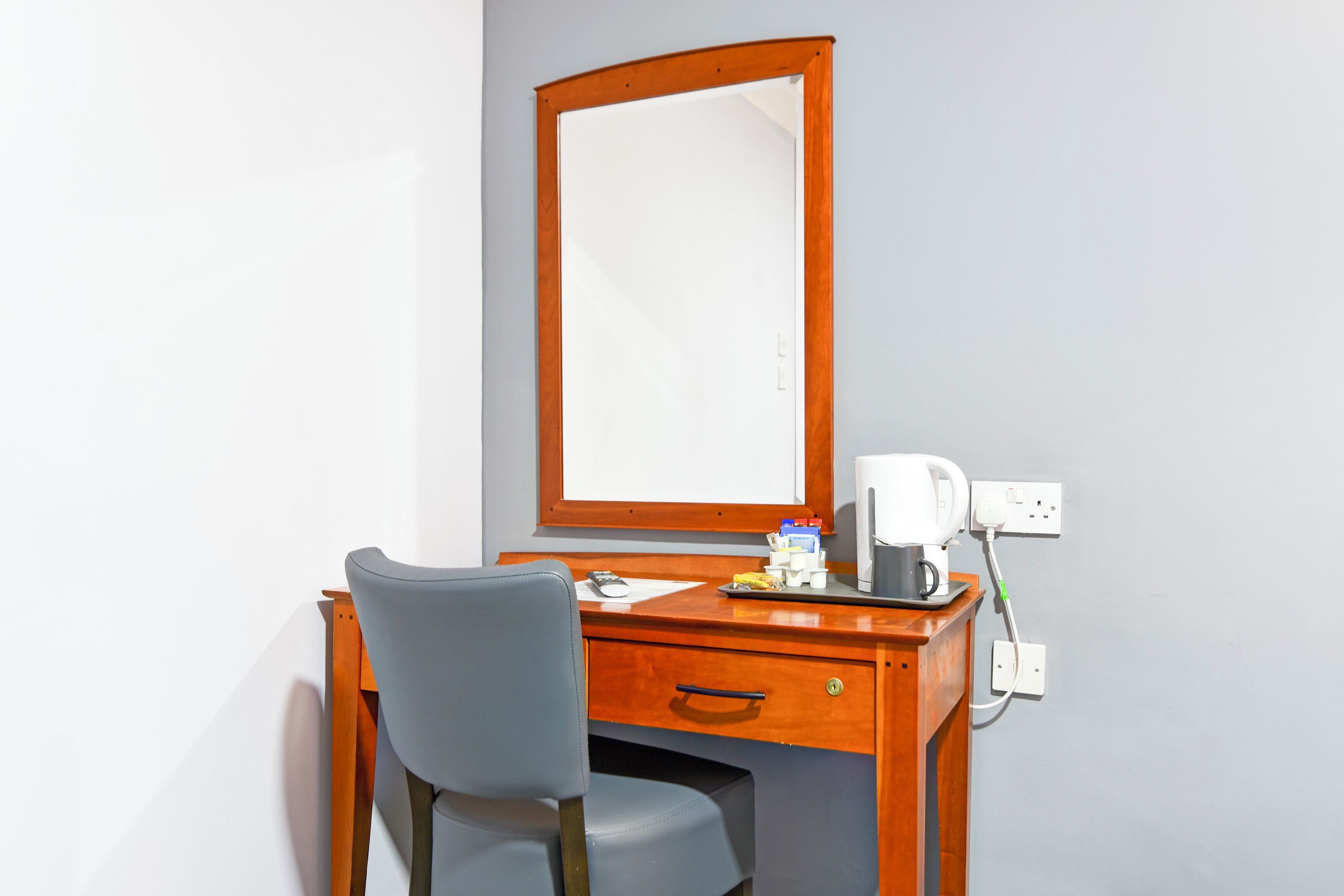 A wooden desk with a chair, a mirror, a coffee maker, and a power outlet.