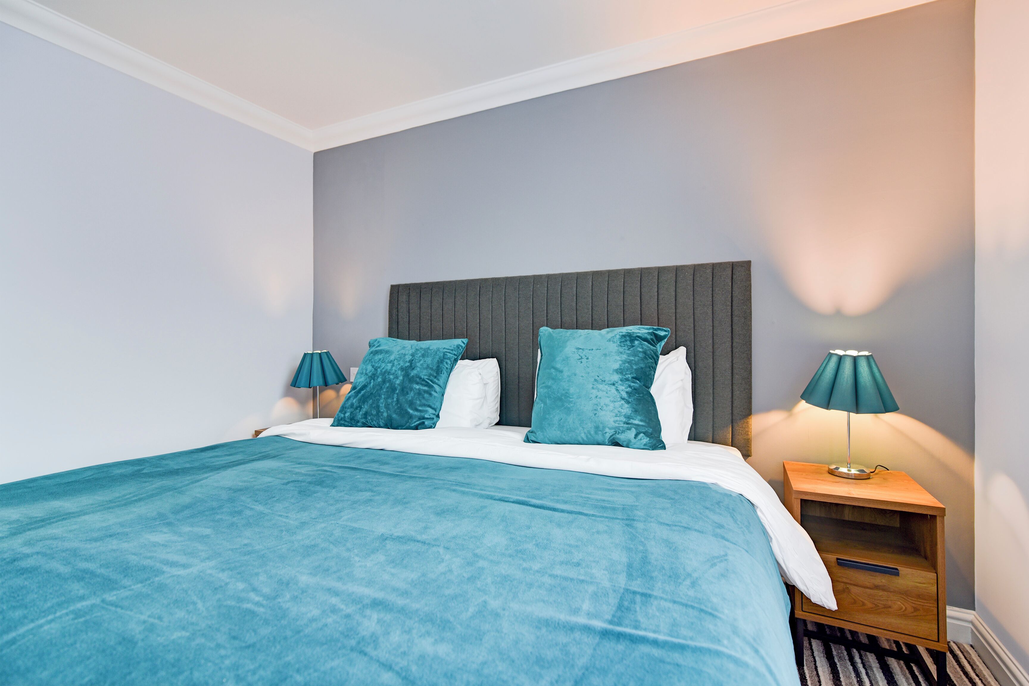 A neatly made bed with a teal bedspread and matching pillows, a wooden nightstand with a lamp, and a grey headboard.
