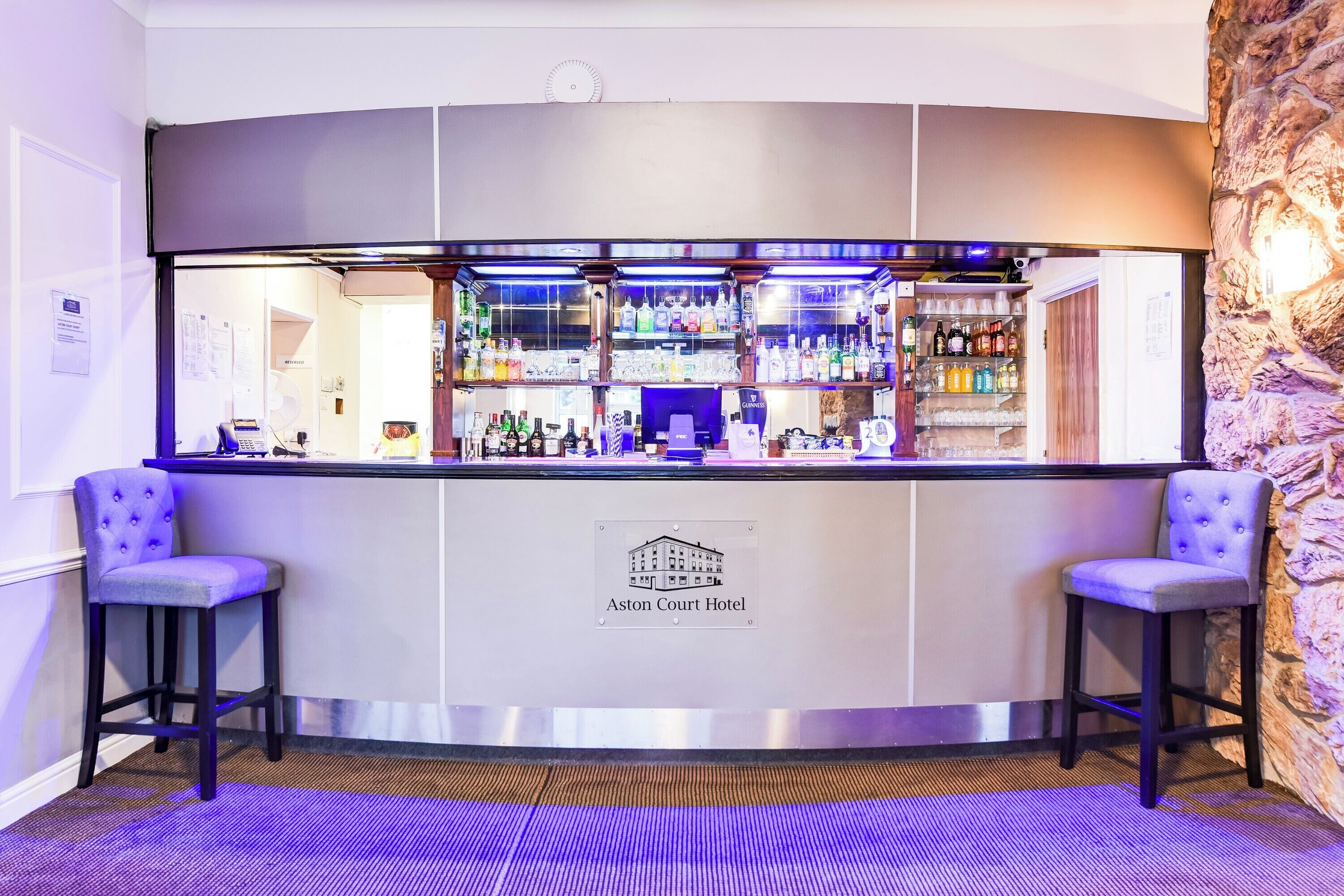 Un coin bar avec un mur en pierre, deux tabourets et une enseigne indiquant qu’il fait partie de l’hôtel Aston Court.