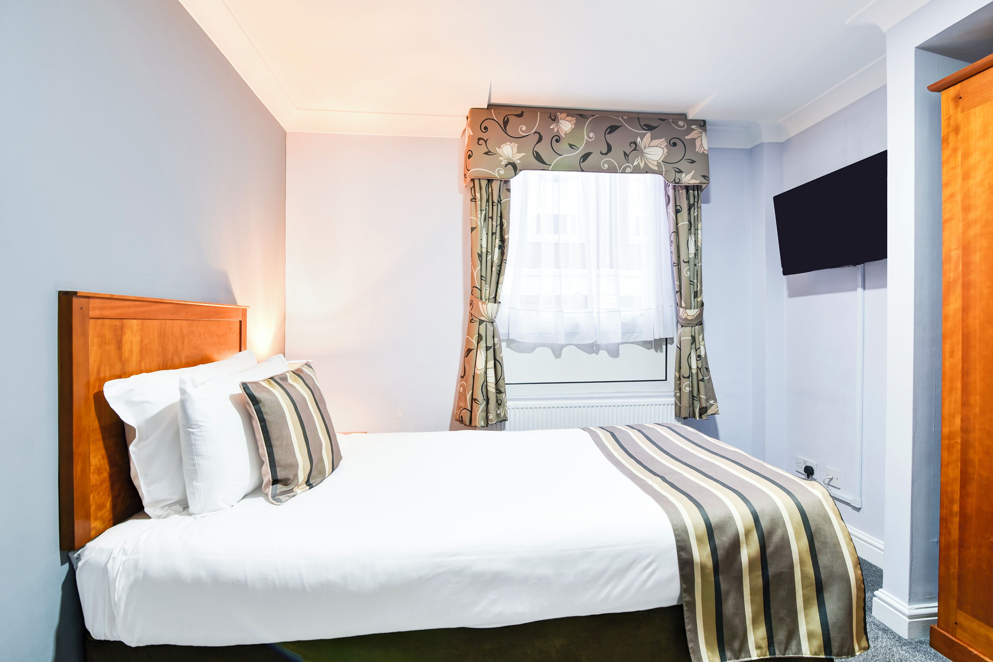 Un lit bien fait, avec une tête de lit en bois, une fenêtre ornée de rideaux à motifs et un téléviseur à écran plat fixé au mur.