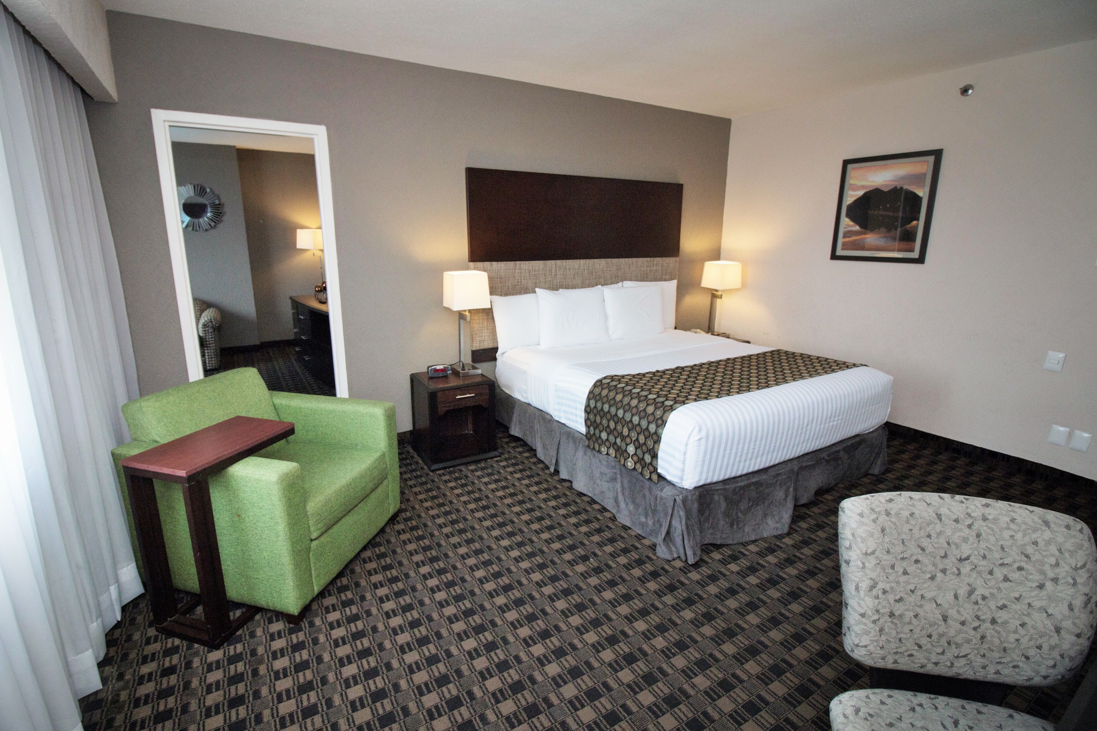 A hotel room with a bed, a green armchair, a small table, a patterned carpet, and a framed picture on the wall.