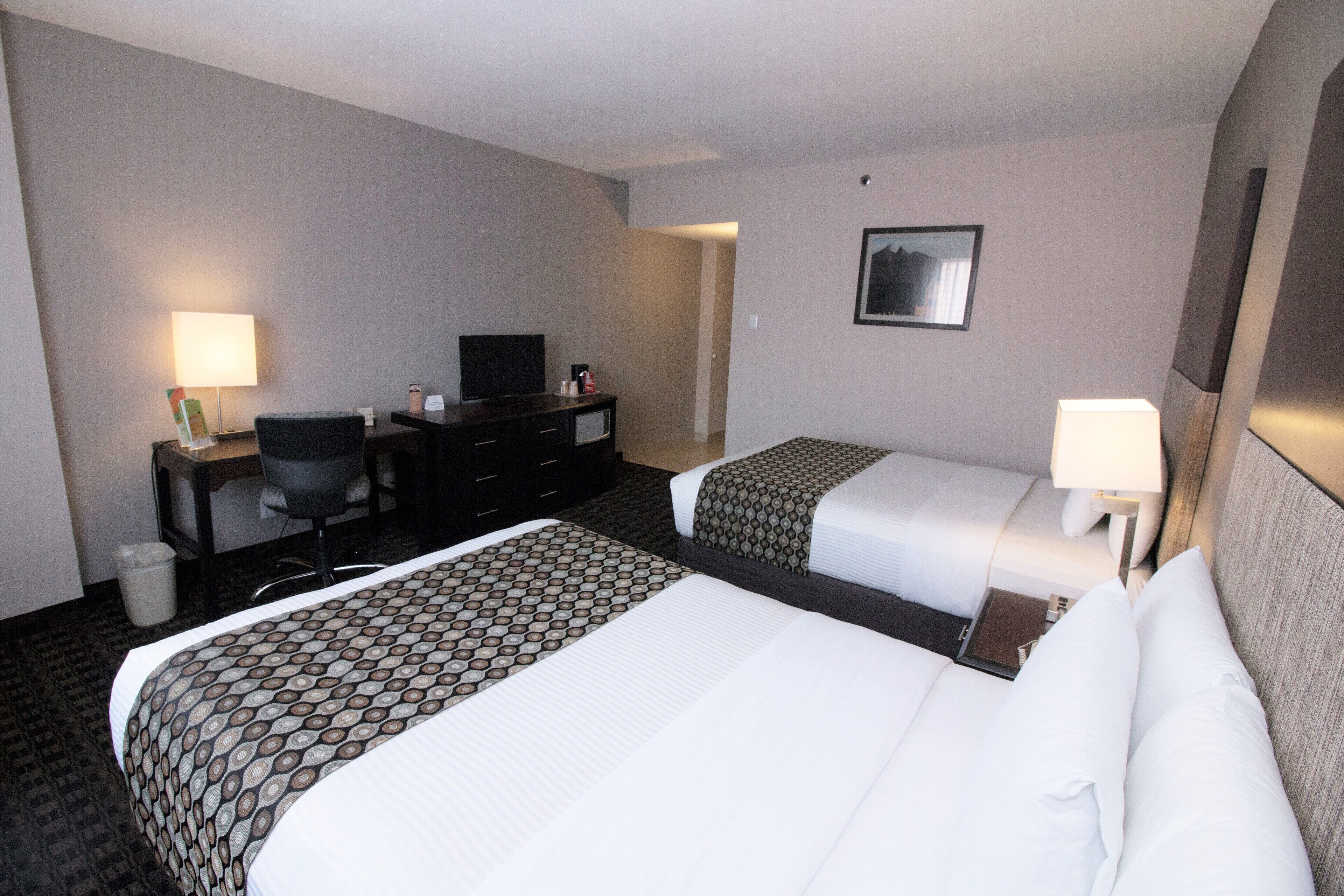Quarto de hotel com duas camas, uma mesa de trabalho com cadeira, televisão e lixeira.