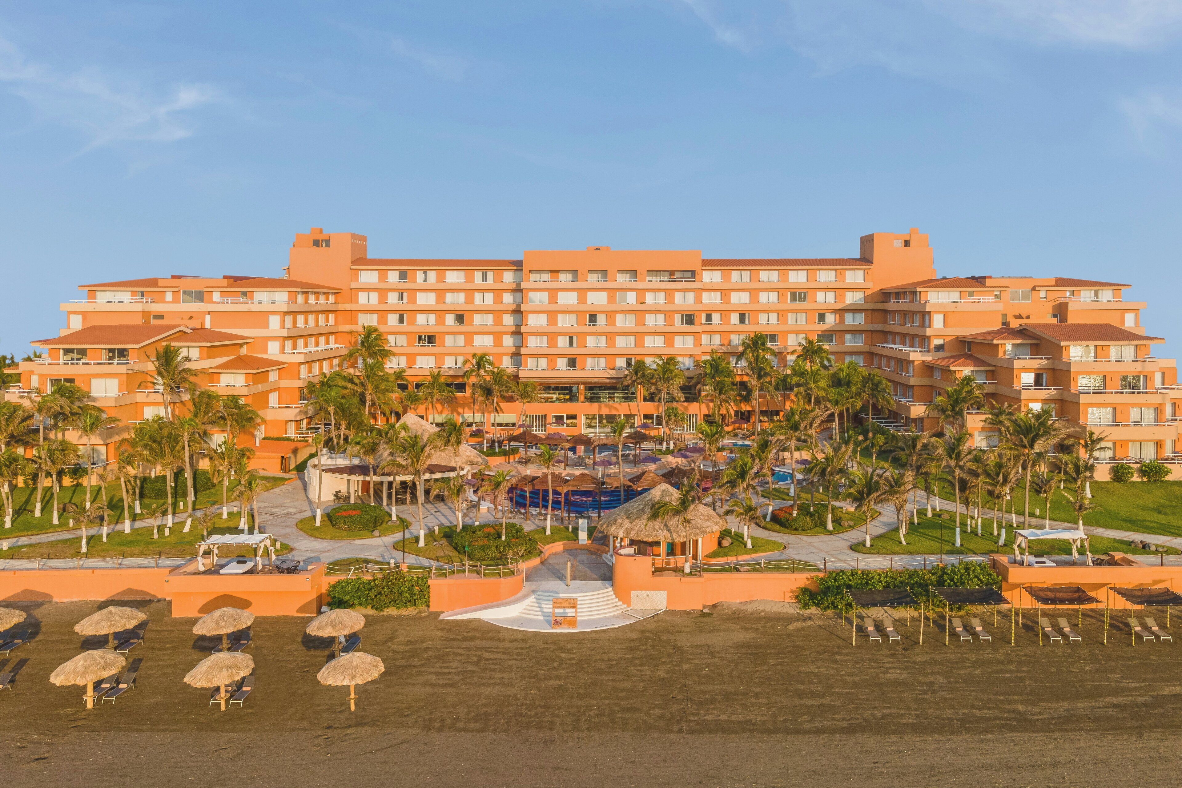 A large hotel with multiple floors, a central courtyard, and a pool area surrounded by palm trees.