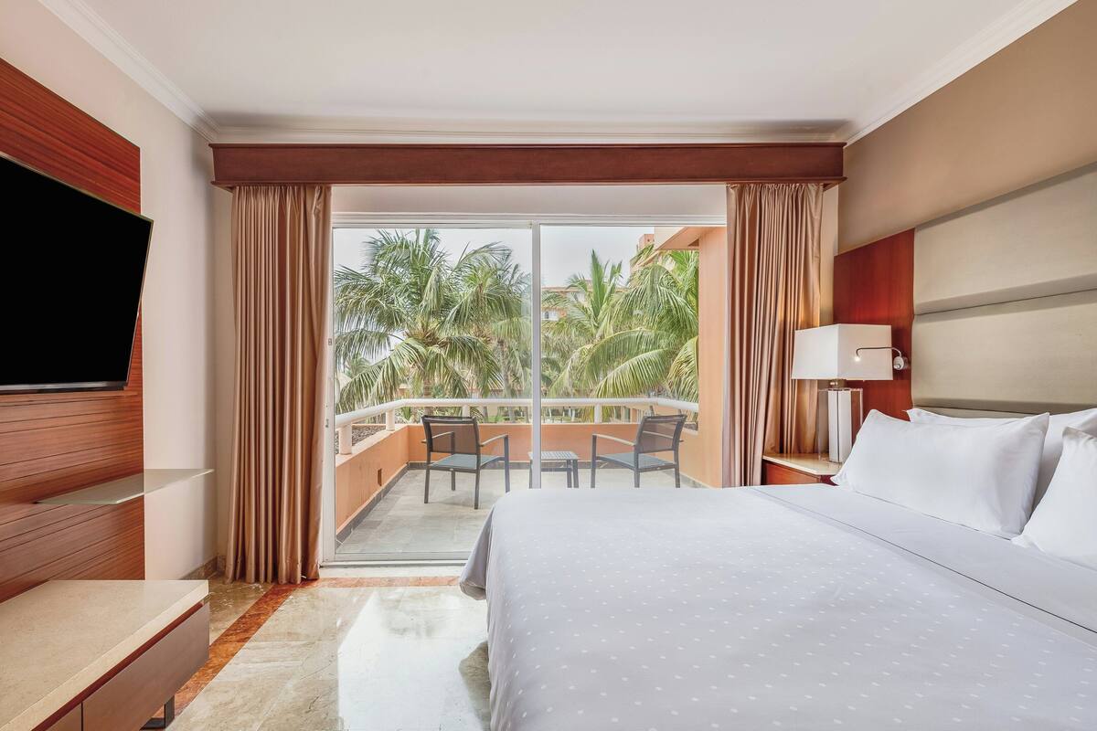 A hotel room with a bed, a TV, a balcony with a table and chairs, and a view of palm trees.