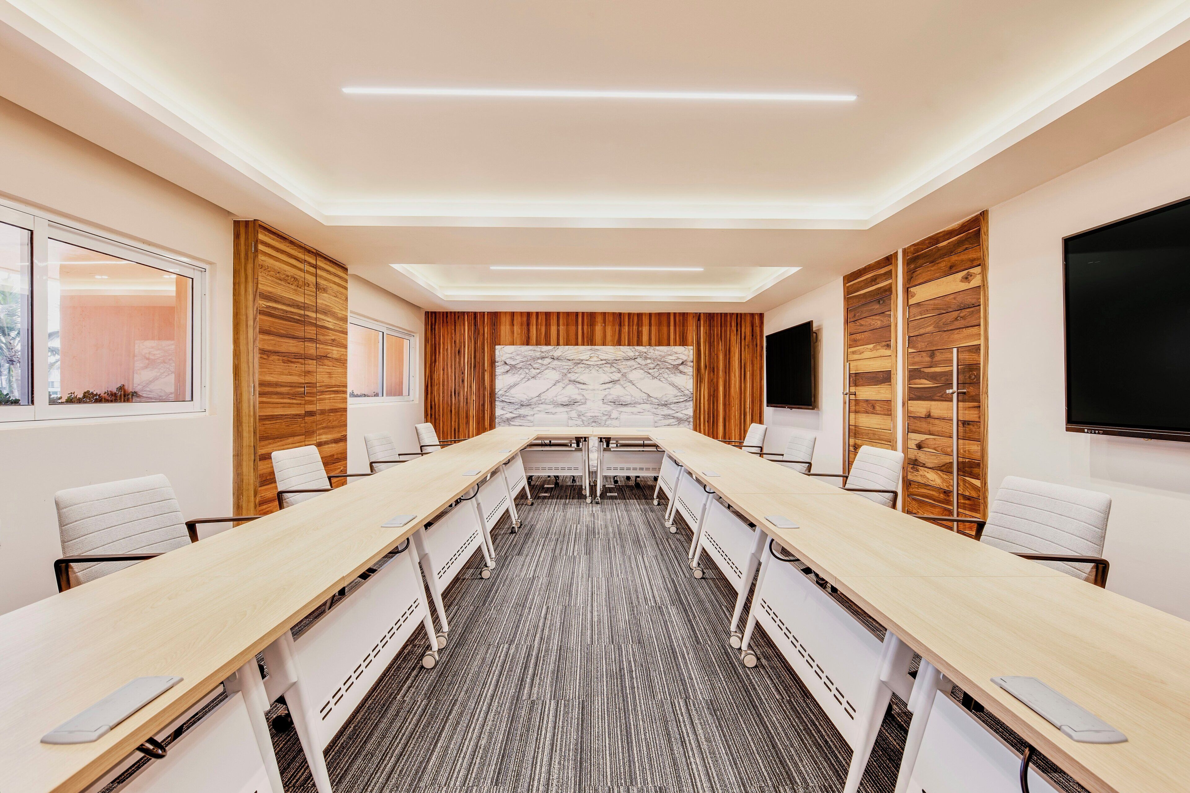 A conference room with a long table, white chairs, a marble accent wall, wooden paneling, and a flat-screen TV.