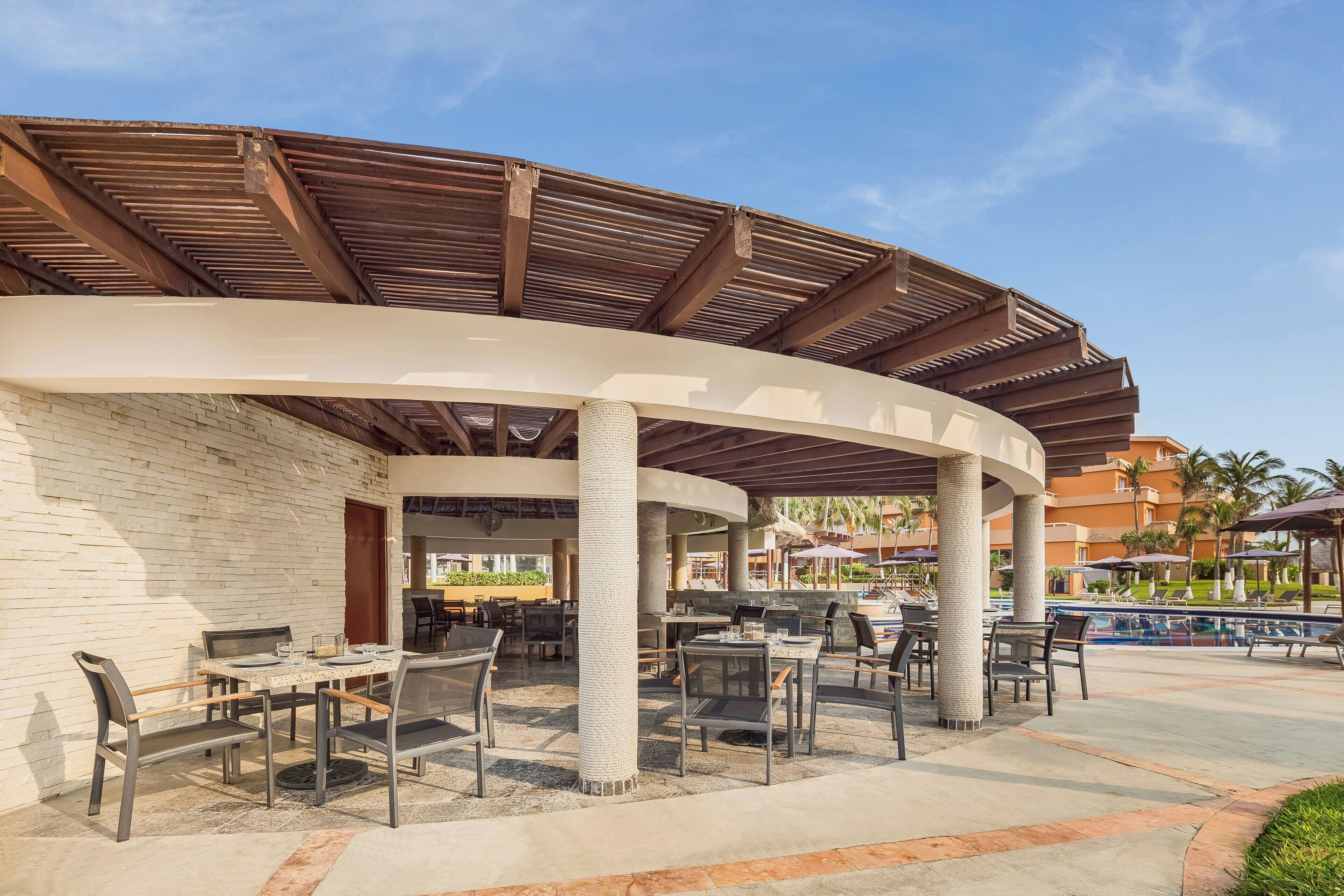 An outdoor patio area with tables and chairs, a pool, and a covered seating area with a curved roof.