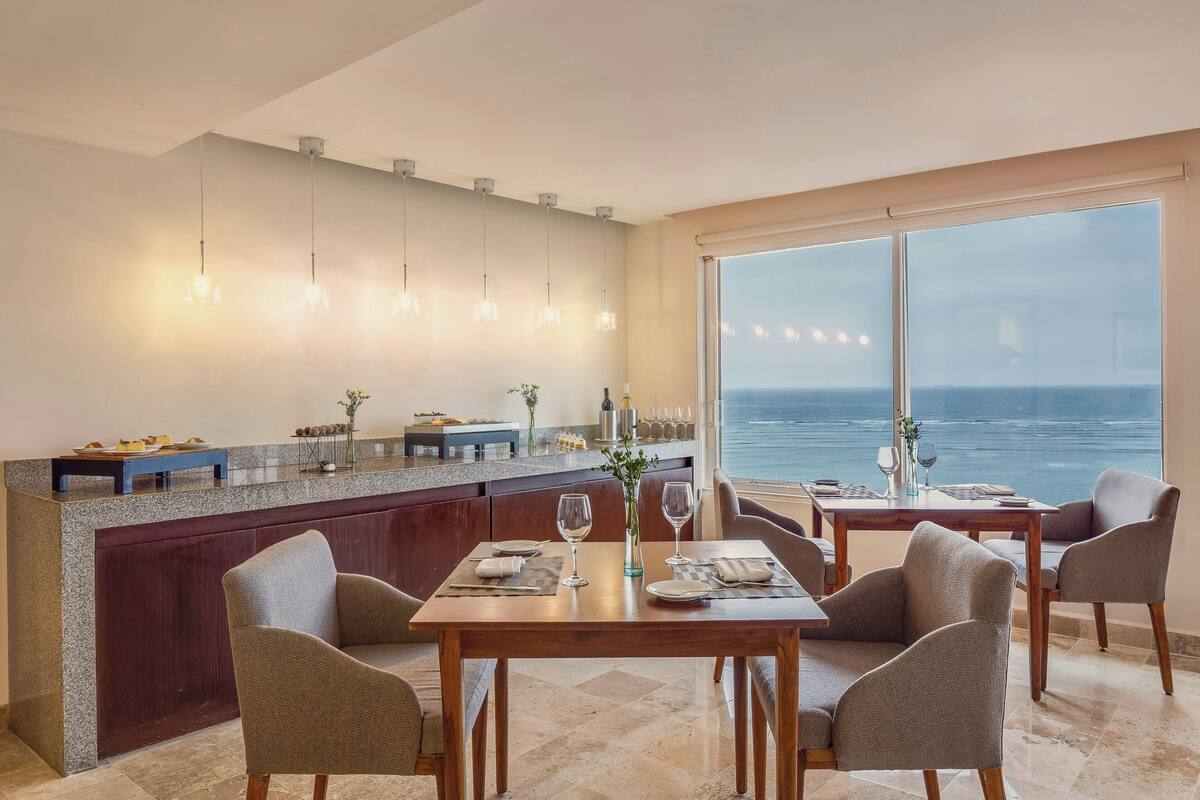 A dining area with a view of the ocean, a bar with a stone countertop, and modern pendant lighting.