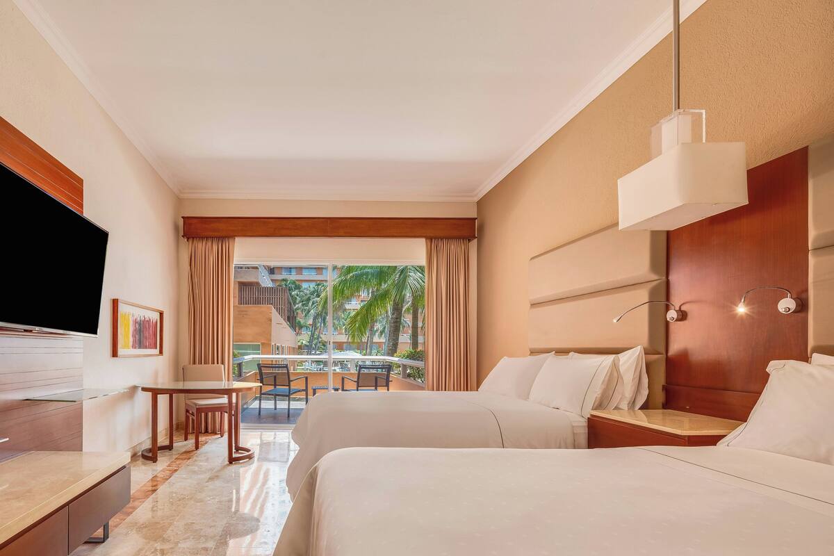 A hotel room with two beds, a TV, a desk, and a view of palm trees.