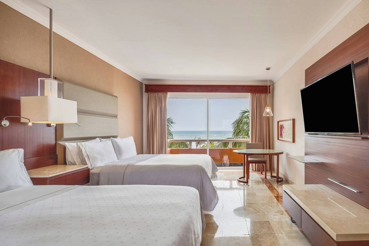 A hotel room with two beds, a TV, a balcony with a table, and a view of the ocean.