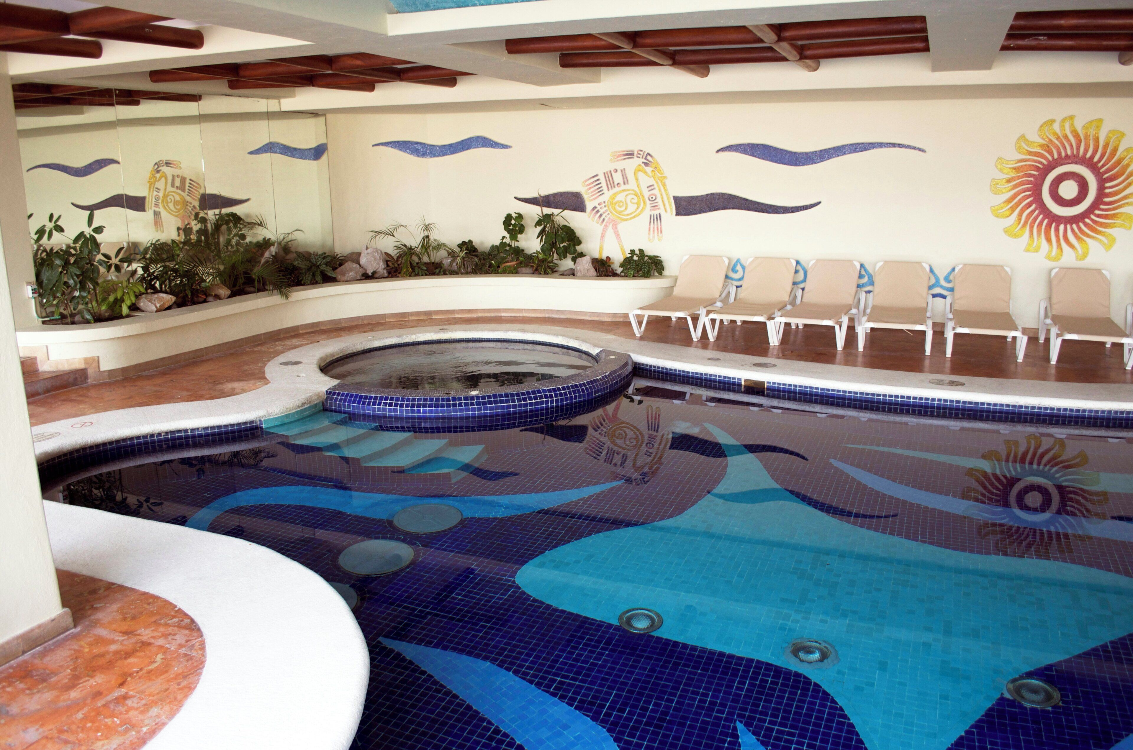 Una piscina con un mural de sol, sillas de descanso y plantas tropicales.