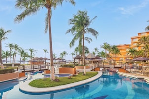 Zona de piscina en un resort, con palmeras, sillas de descanso y un edificio al fondo.