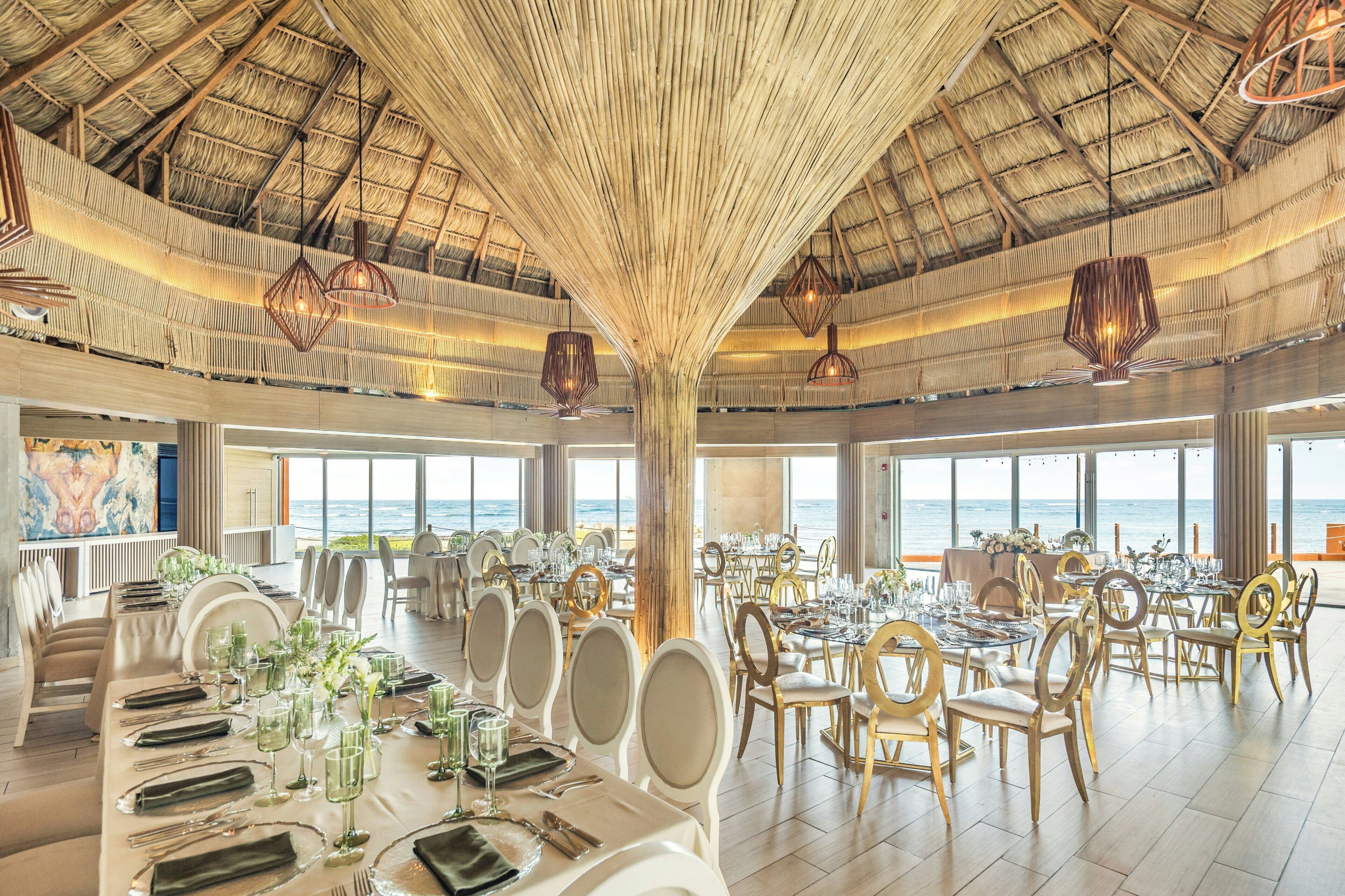 A spacious dining area with multiple tables set for a meal, large windows offering a view of the sea, and a high ceiling with a thatched roof design.