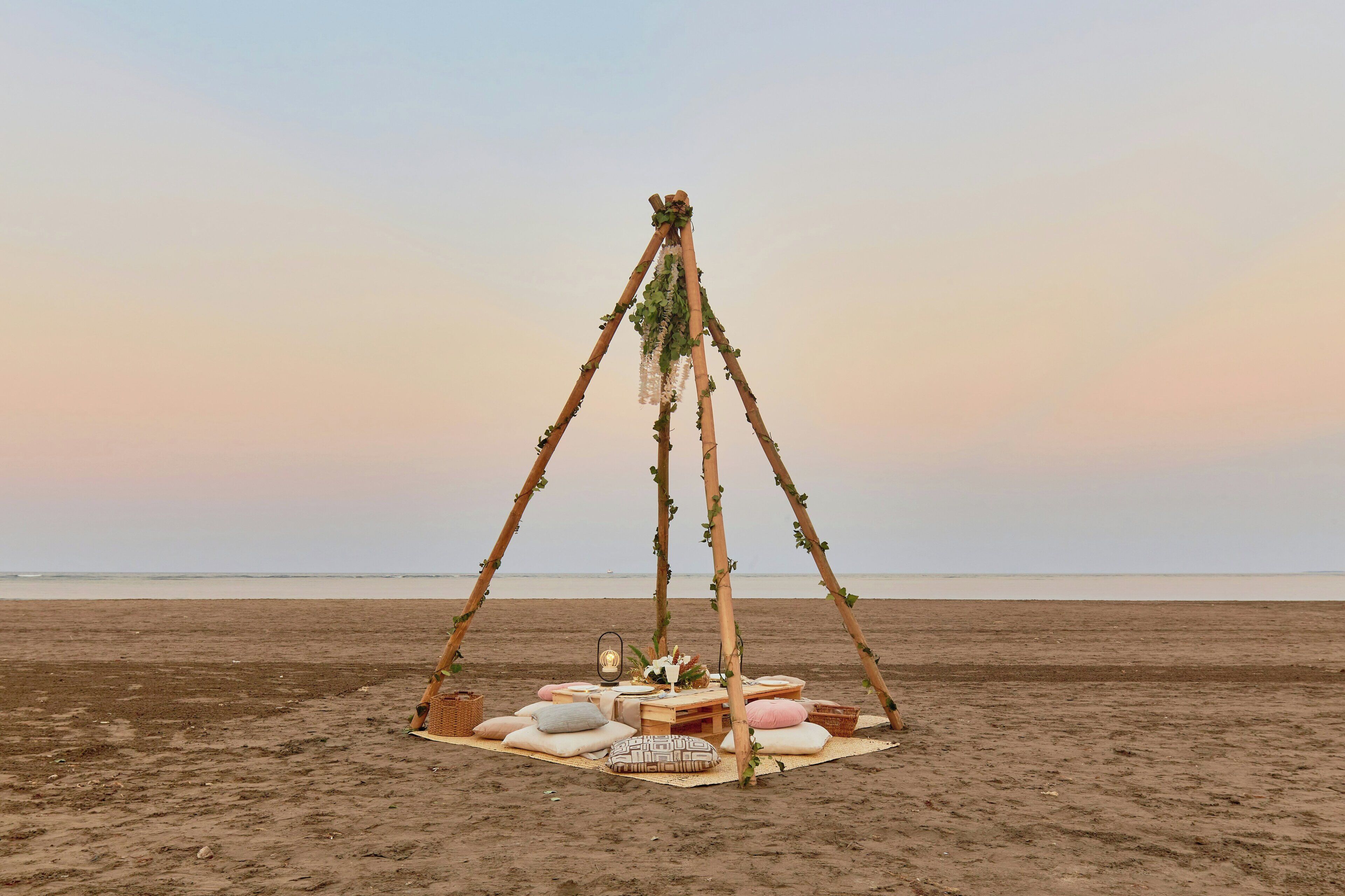 A beach setting with a wooden structure adorned with greenery, a picnic setup with cushions and a table, and a view of the ocean.