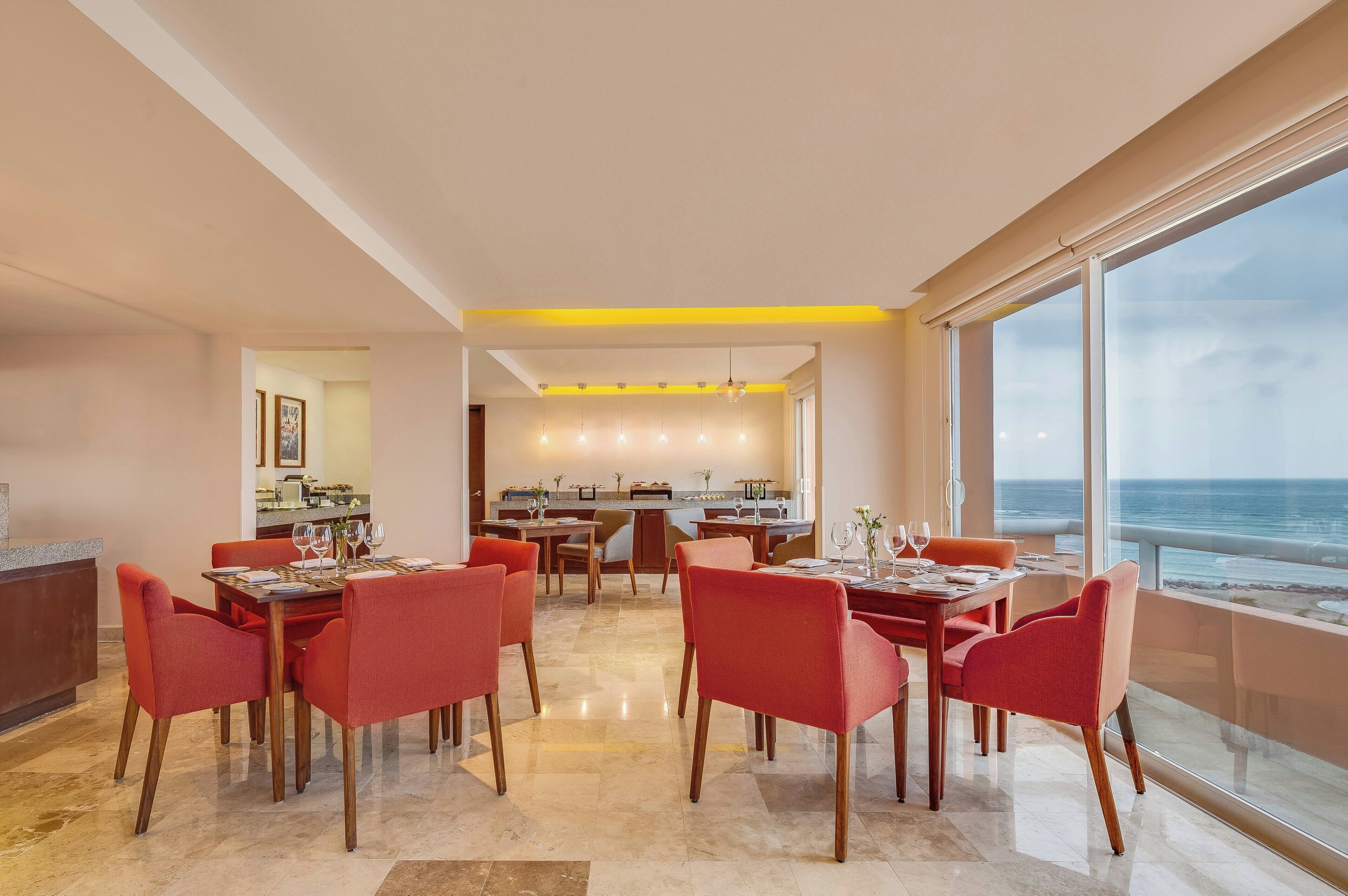 A spacious dining area with red chairs and a central table, a kitchen in the background, and a large window with a view of the ocean.