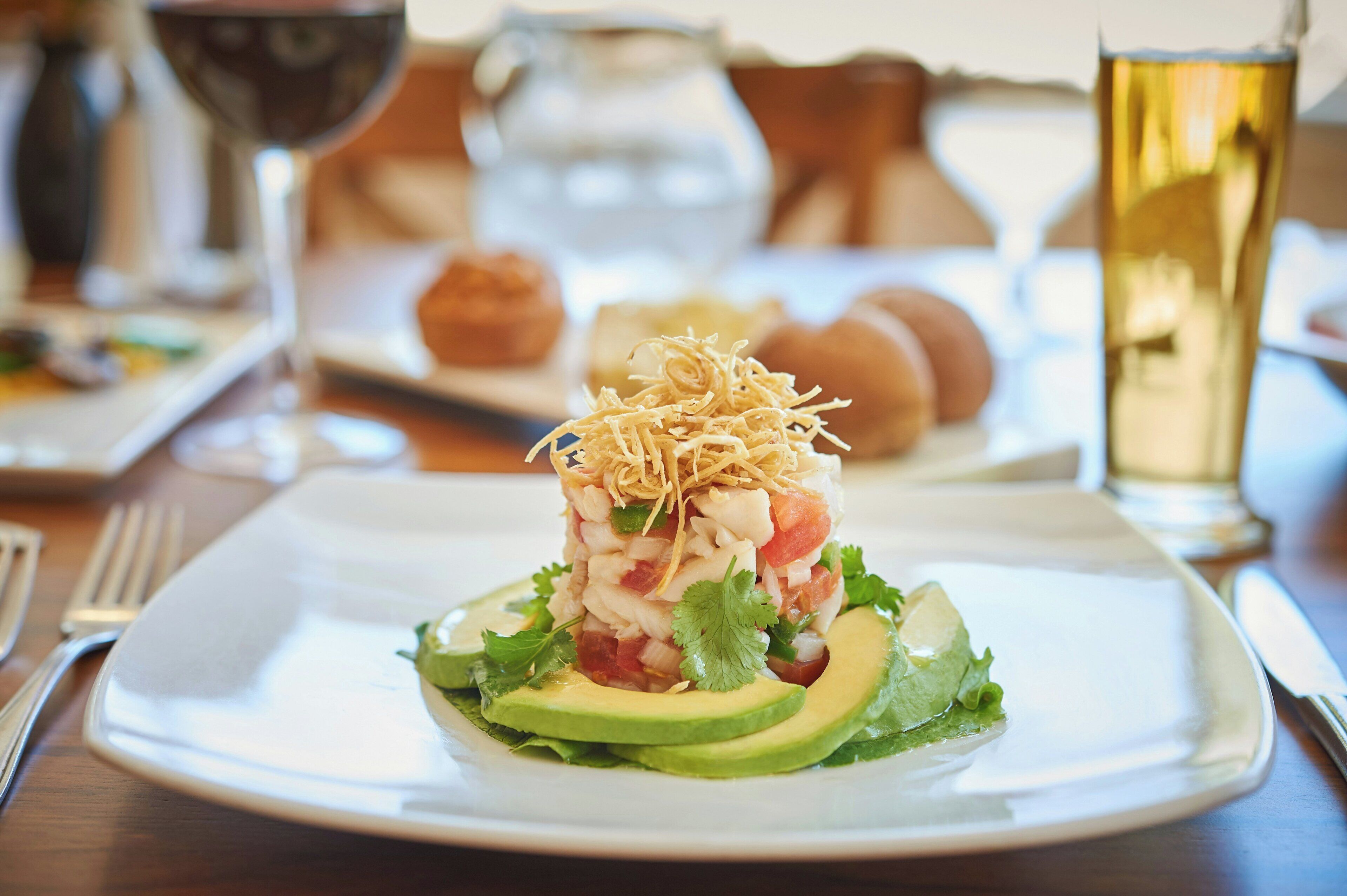 A plate of seafood salad with avocado and shredded cheese, accompanied by a glass of white wine and a pitcher of water on a wooden table.