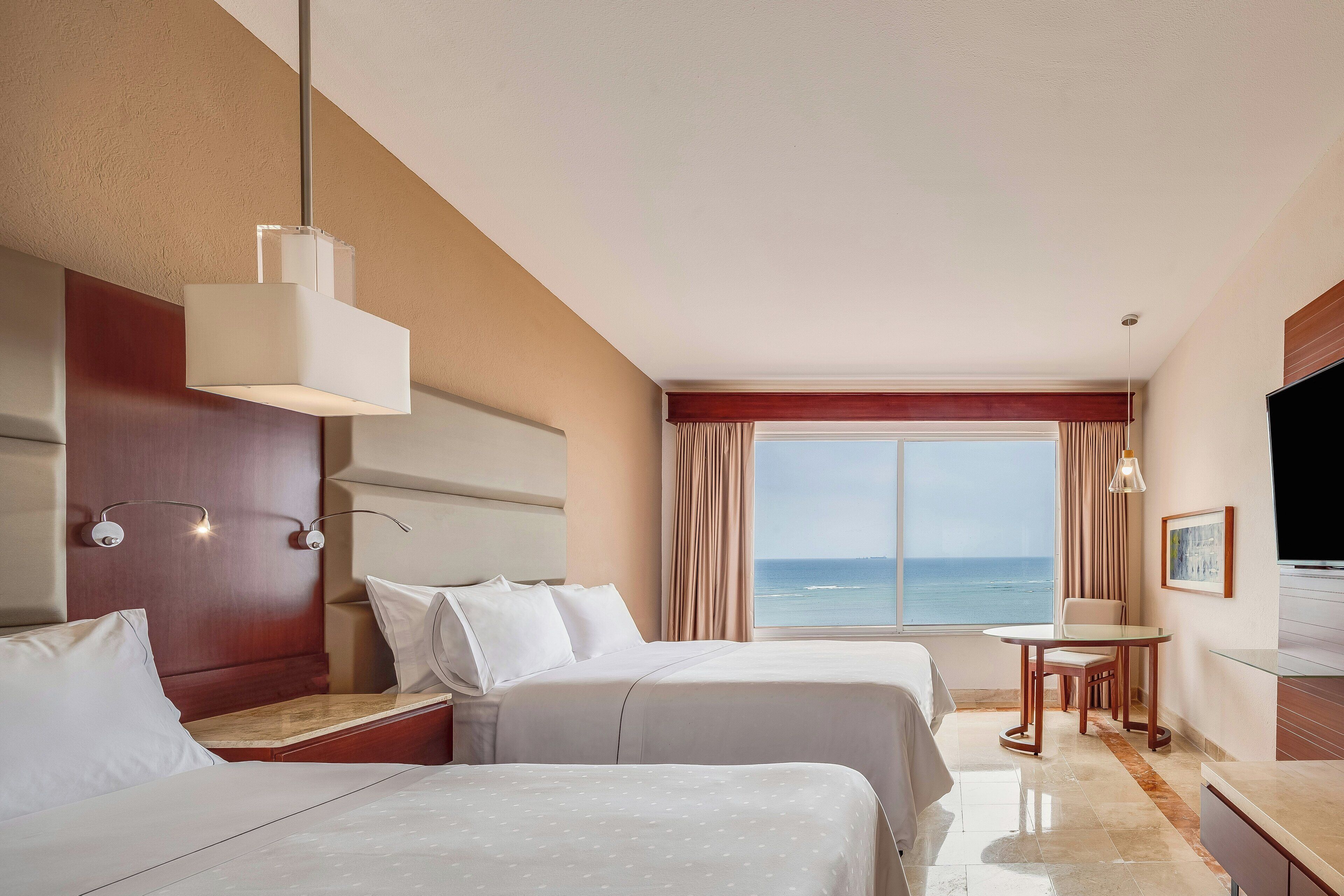 A hotel room with two beds, a dining area, a TV, and a view of the ocean.