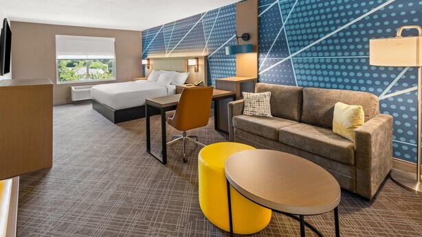 Una habitación de hotel con cama, sofá, otoman amarillo, mesa redonda, escritorio y silla.
