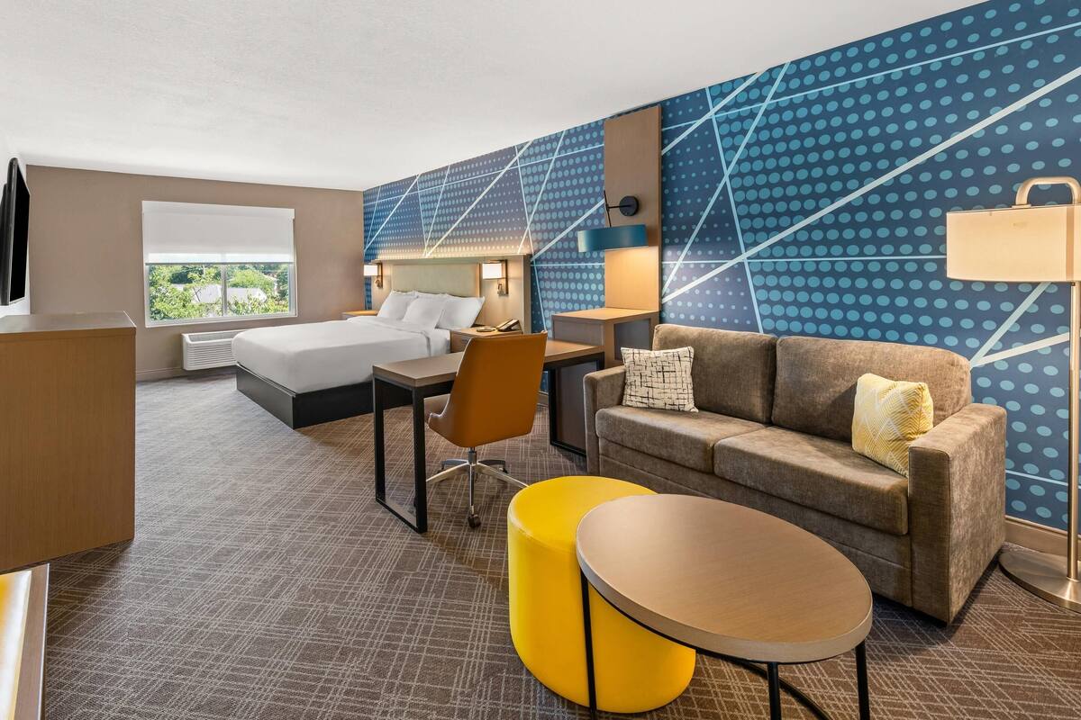 A hotel room with a bed, a sofa, a yellow ottoman, a round table, a desk, and a chair.