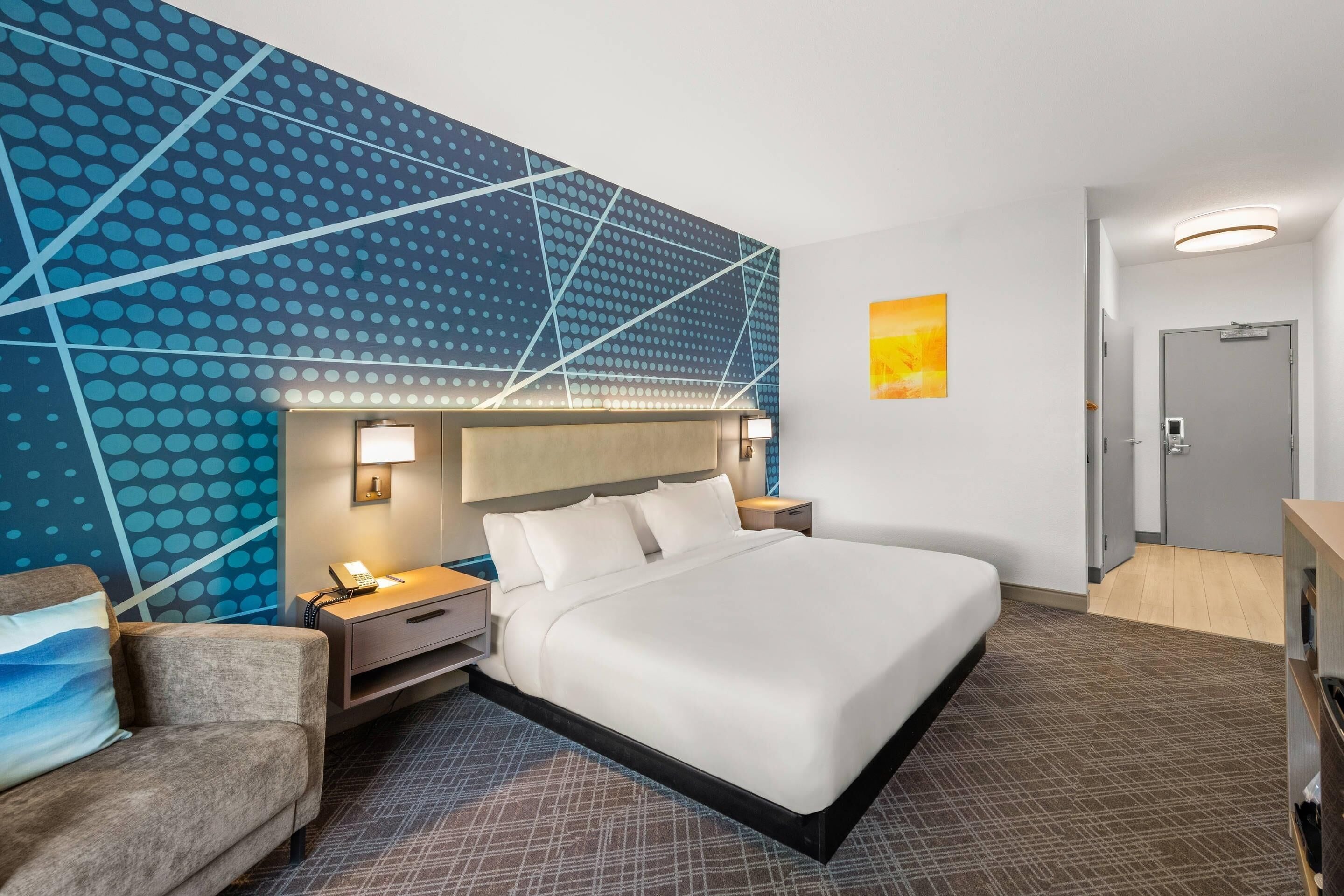 A hotel room with a large bed, a desk, a chair, a nightstand, a wall with a geometric pattern, and a door.