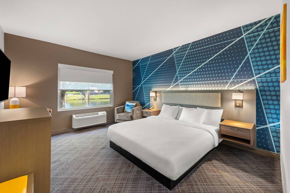 A hotel room with a large bed, a desk, a chair, a window with a view of a park, and a wall with a geometric pattern.