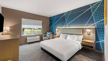 A hotel room with a large bed, a desk, a chair, a window with a view of a park, and a wall with a geometric pattern.