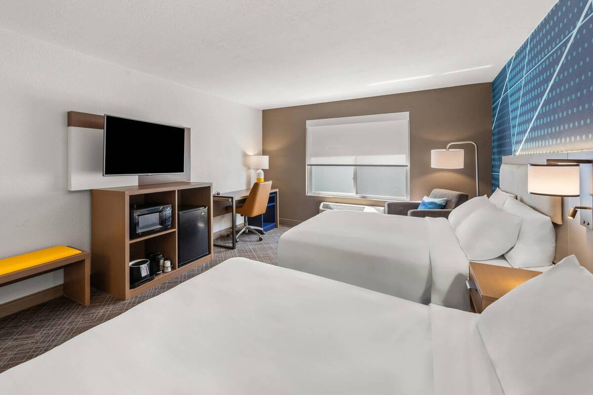A hotel room with two beds, a TV, a desk with a chair, a microwave, a coffee maker, and a large window.