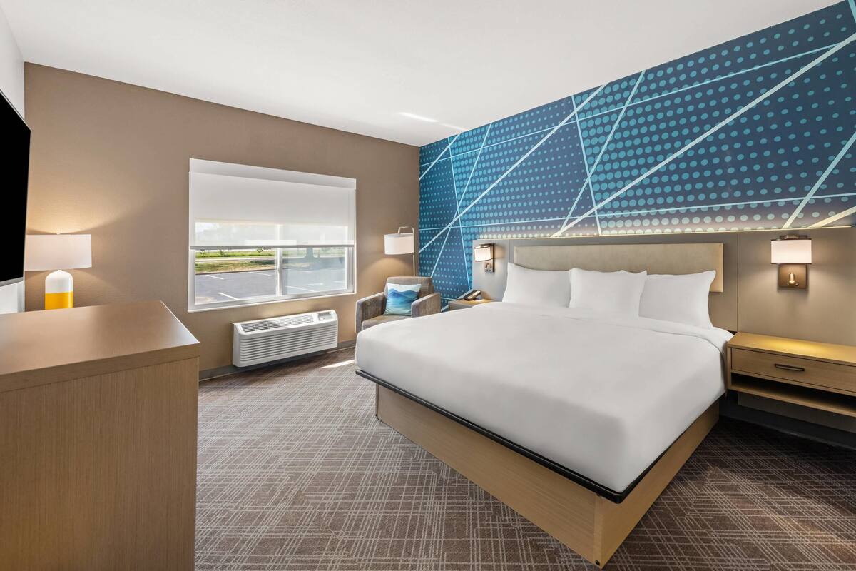 A modern hotel room with a large bed, a desk, a chair, a TV, and a window with a view of the outdoors.