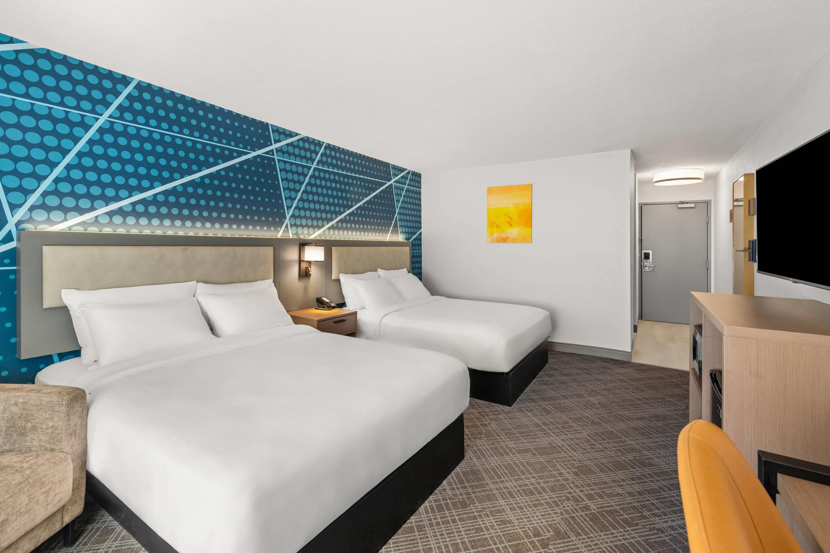 A hotel room with two beds, a desk, a chair, a TV, and a wall with a geometric pattern.