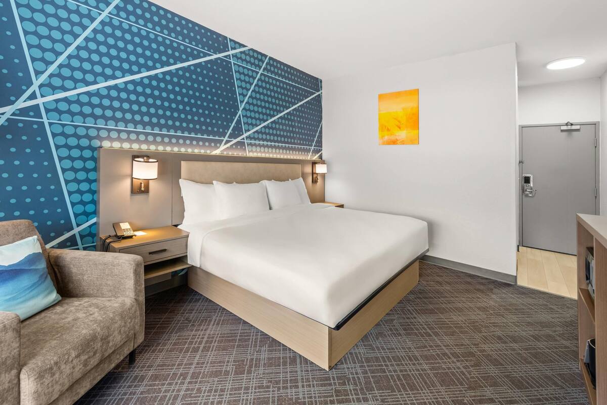 A hotel room with a bed, nightstands, a chair, a wall with a geometric pattern, and a door.